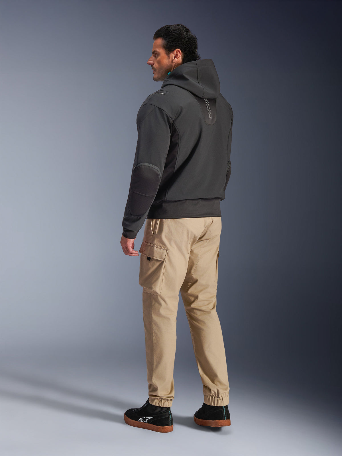 Alpinestars Moblast Waterdichte Jas, zwarte motor tech hoodie, zij- en achteraanzicht, gedragen met beige cargo broek en zwarte sneakers, voorzien van een ontwerp met capuchon, technische textielconstructie en een moderne stedelijke esthetiek.