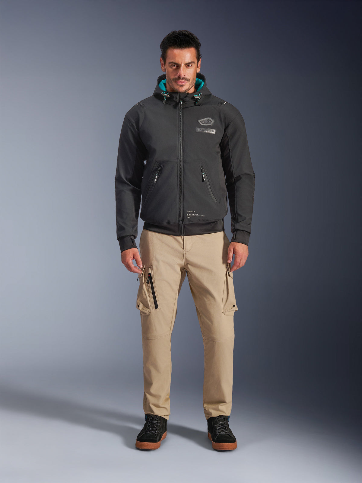 Alpinestars Moblast Waterproof Jacket, zwarte urban motorhoodie met groenblauwe binnenvoering in de capuchon, gedragen met een beige cargobroek en zwarte hoge boots, eigentijdse all-season rijkleding