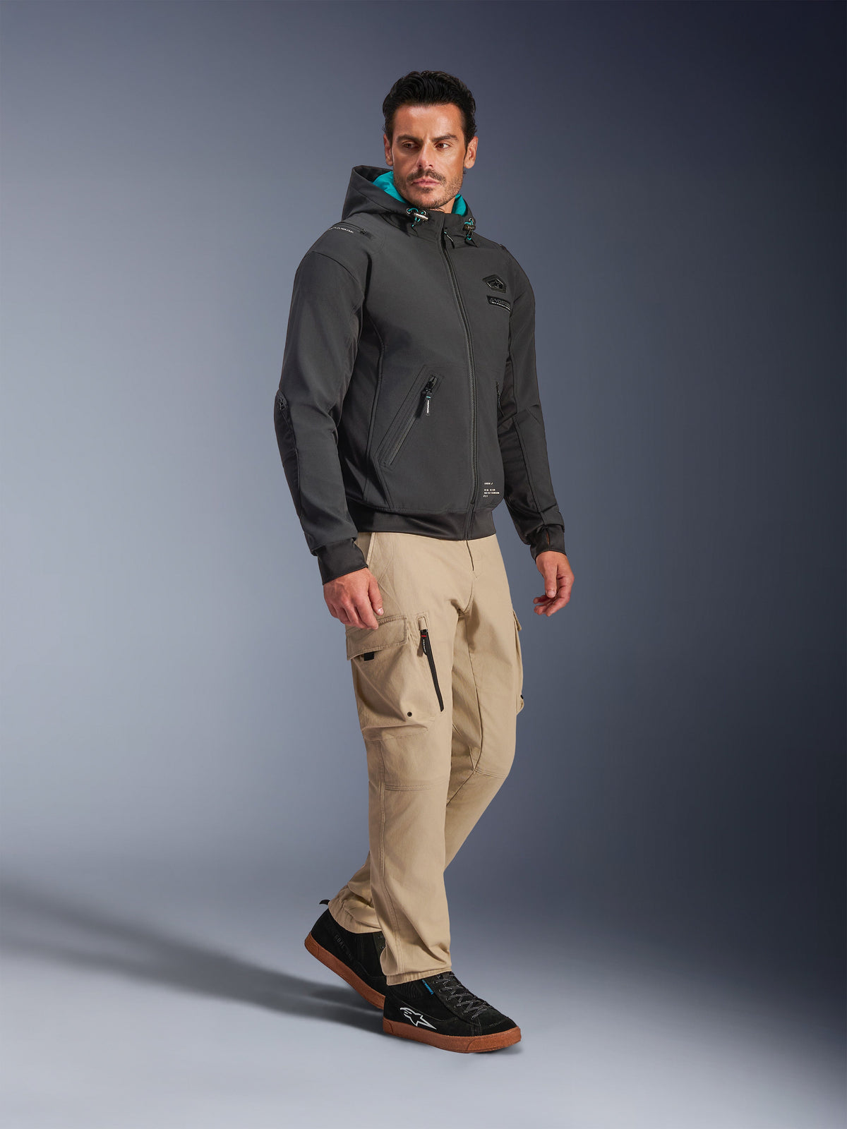 Mannelijk model draagt Alpinestars Moblast Waterproof Jas in het zwart, stedelijke motorhoodie met blauwgroen gevoerde capuchon, gecombineerd met een kaki cargobroek en zwarte sneakers, modern minimalistisch ontwerp met technische stretchstof voor woon-werkverkeer in alle weersomstandigheden