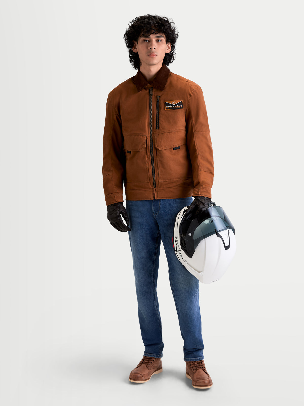 Alpinestars Mustang WP jas, urban contemporary heren motorjas in donkerroest en donkerbruin, vooraanzicht van een model in werkkleding-stijl rijkleding met een witte helm