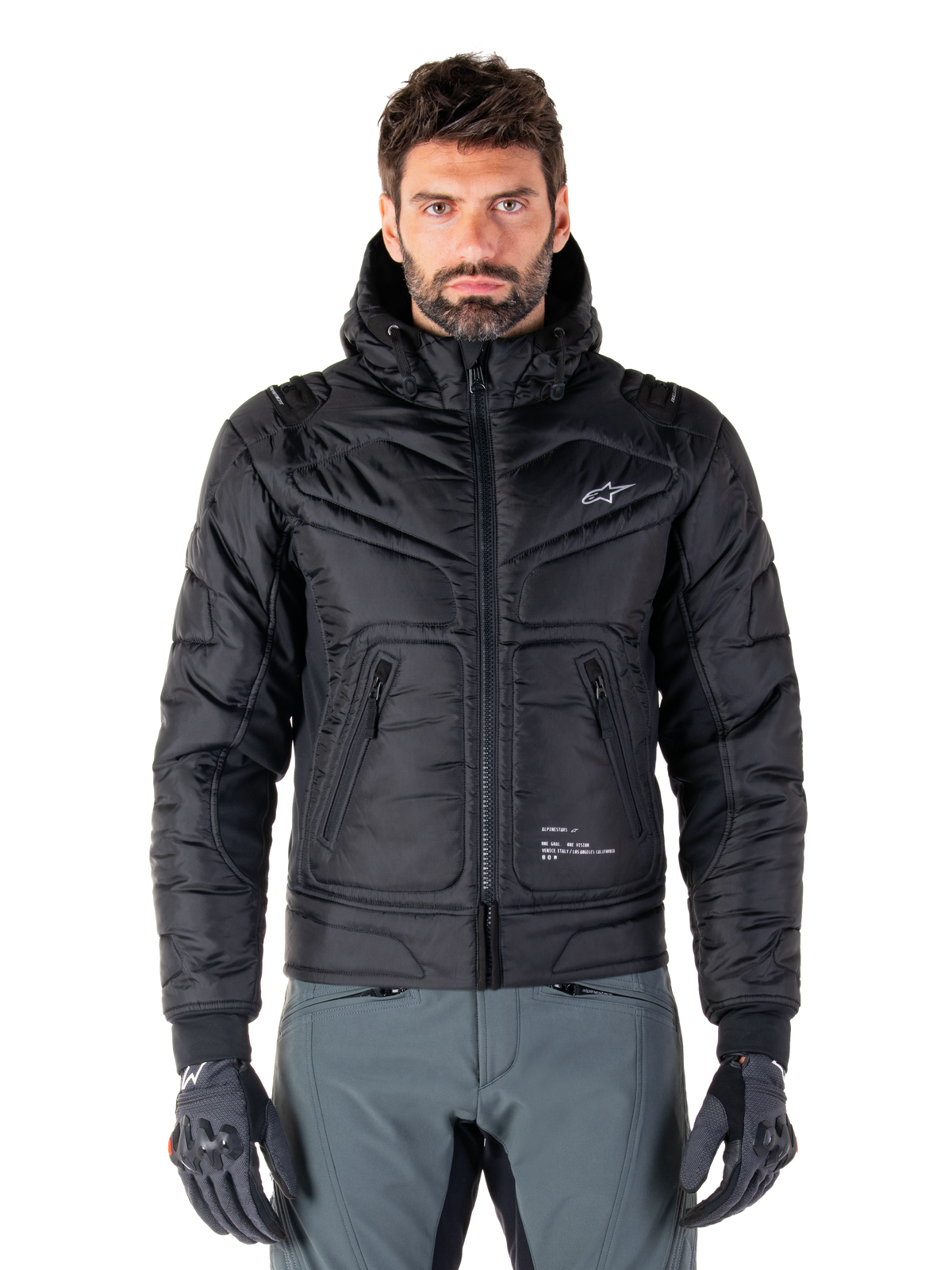 Alpinestars Mohobbs Waterproof Jacket, zwarte gewatteerde bomber motorjas, gewatteerde poly-stof met ritssluiting en ritszakken, klein wit logo op de borst, gedragen door een model met grijze broek en handschoenen, urban streetwear-ontwerp voor rijden in de stad