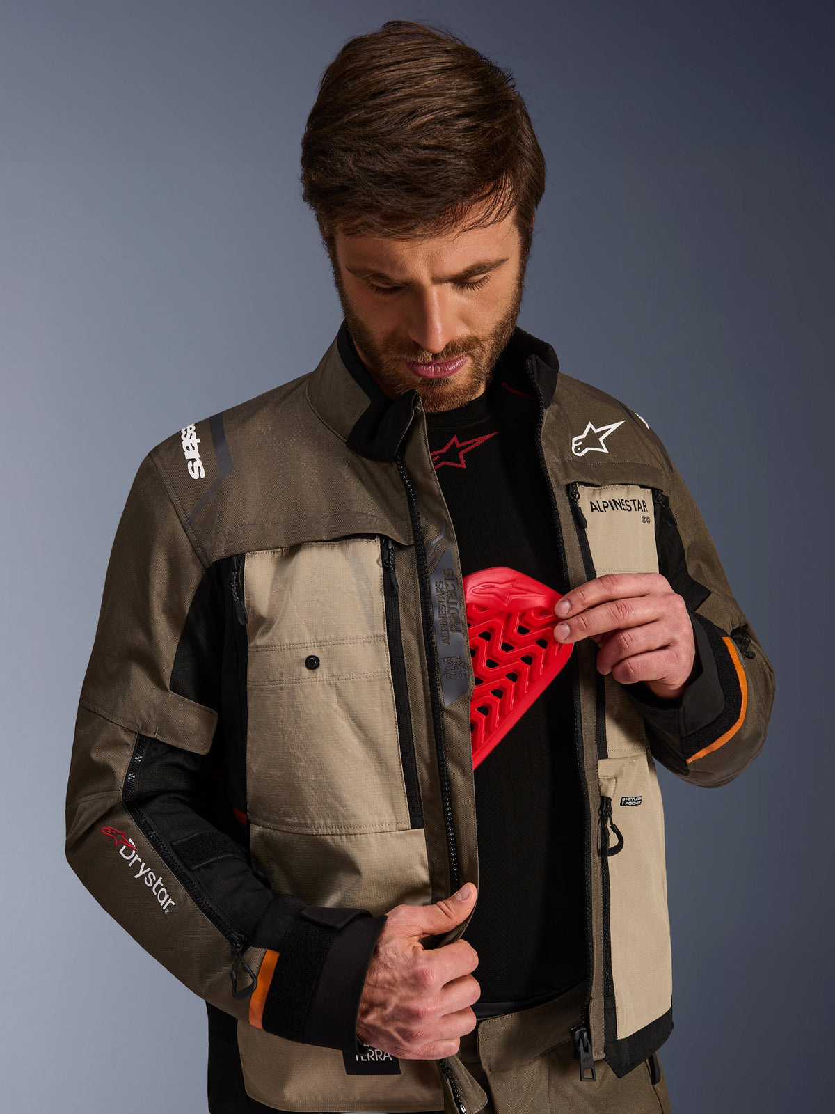 Alpinestars Cusco Drystar® Jacket, Walnut Aluminum Black, worn by a man demonstrating the chest protector pocket with a red armor insert, textile motorcycle jacket with zippered vents and branding for adventure touring.