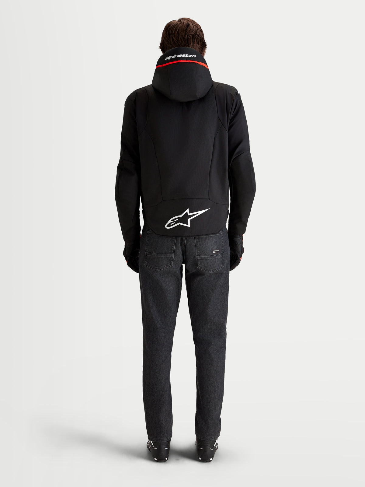 Alpinestars Zaca V 2 Waterproof Jas, heren waterdichte motorjas van achteren bekeken, met geïntegreerde capuchon, verlengde achterzoen met een wit Astar-logo en donkere denim jeans