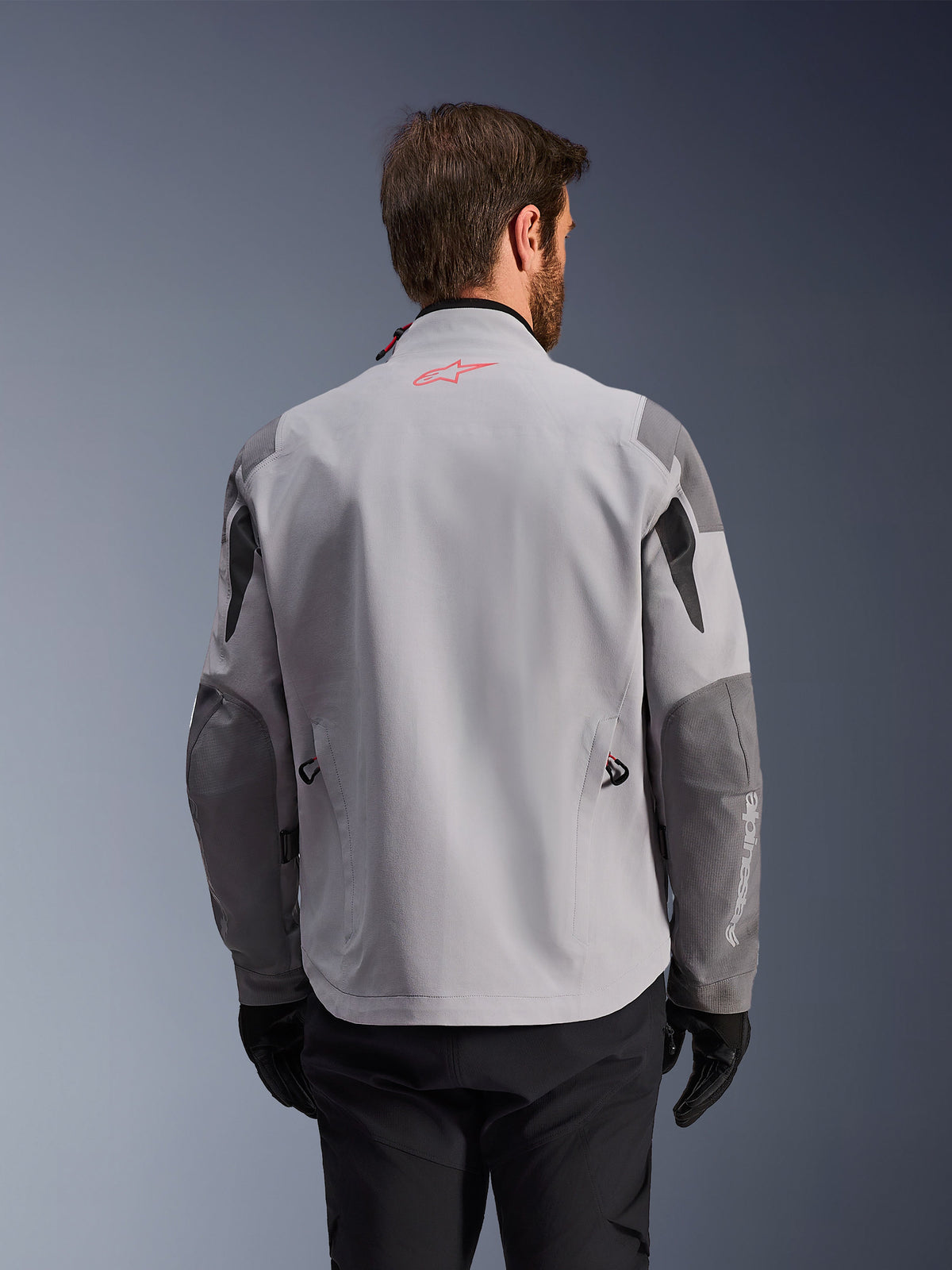 Alpinestars Maxdura Drystar®XF Laminated Armored Shell, achteraanzicht van een adventure motorjas, Mid Gray en Dark Gray, met een rood ster-logo op de kraag en witte verticale merknaam op de mouw, gedragen met zwarte motorhandschoenen en broek