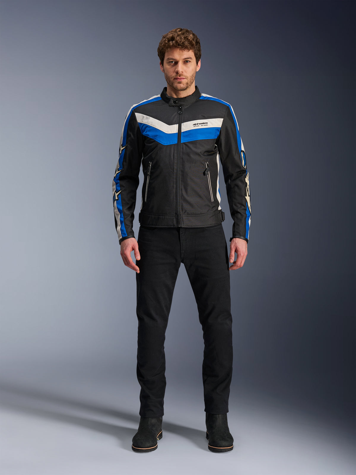 Alpinestars T-Dyno WR Jacket, textiele motorjas, zwart, blauw en ecru, gedragen door een mannelijk model met zwarte broek en laarzen, met chevron-strepen op de borst en erfgoed biker-ontwerp