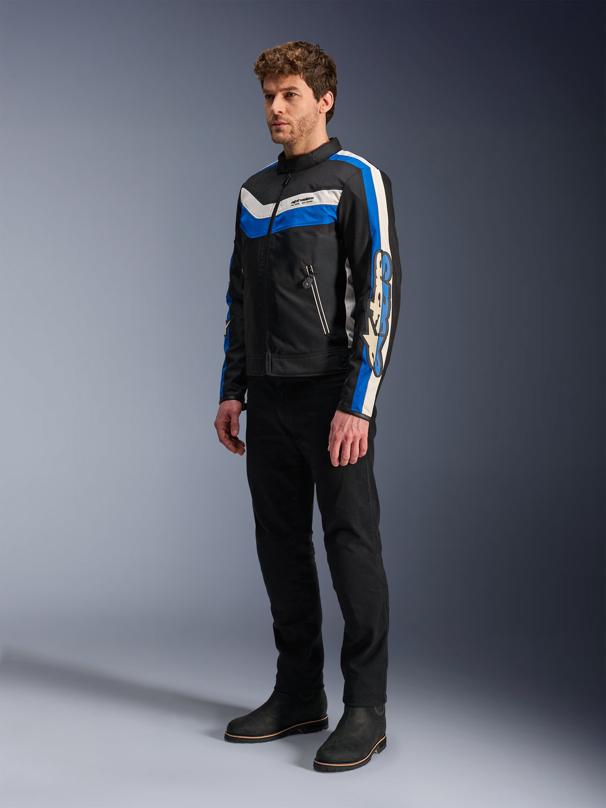Alpinestars T-Dyno WR Jas, textiele motorjas, zwart met blauwe en ecru accenten, vintage-geïnspireerd ontwerp met opvallende strepen en branding op de mouwen, gedragen door model met zwarte broek en laarzen, studiofotografie