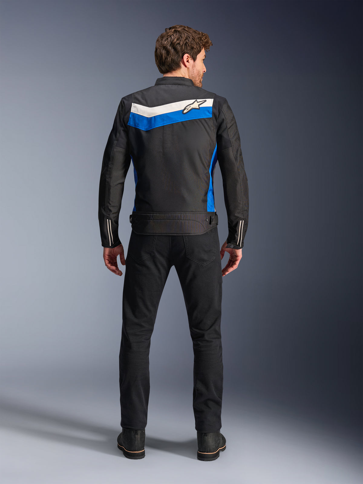 Alpinestars T-Dyno WR Jas, textiele motorjas, kleurstelling Black Blue Ecru, volledig achteraanzicht op model, voorzien van een blauwe en ecru chevron-streep met Alpinestars-logo, gecombineerd met zwarte broek en laarzen, vintage-geïnspireerde urban motorkleding