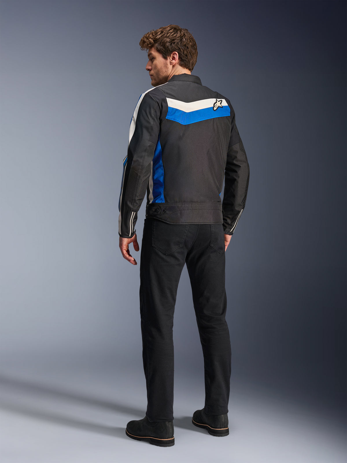 Alpinestars T-Dyno WR jas in Black Blue Ecru, achteraanzicht op een mannelijk model, voorzien van een witte en blauwe chevronstreep over de schouders met een ster-logo, vintage-geïnspireerde textiele motorjas, gedragen met een zwarte broek en laarzen