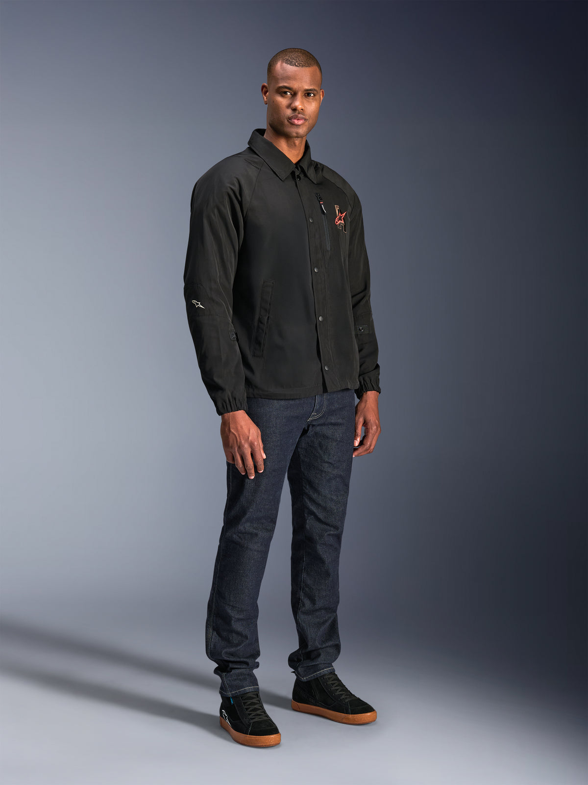 Alpinestars Revved WR Jacket in het zwart, urban motorjas in coaches-stijl gedragen door een model met donkerblauwe jeans en zwarte sneakers, voorzien van een drukknoopsluiting aan de voorzijde en een geborduurd logo, minimalistisch en sportief ontwerp voor rijden op de weg.