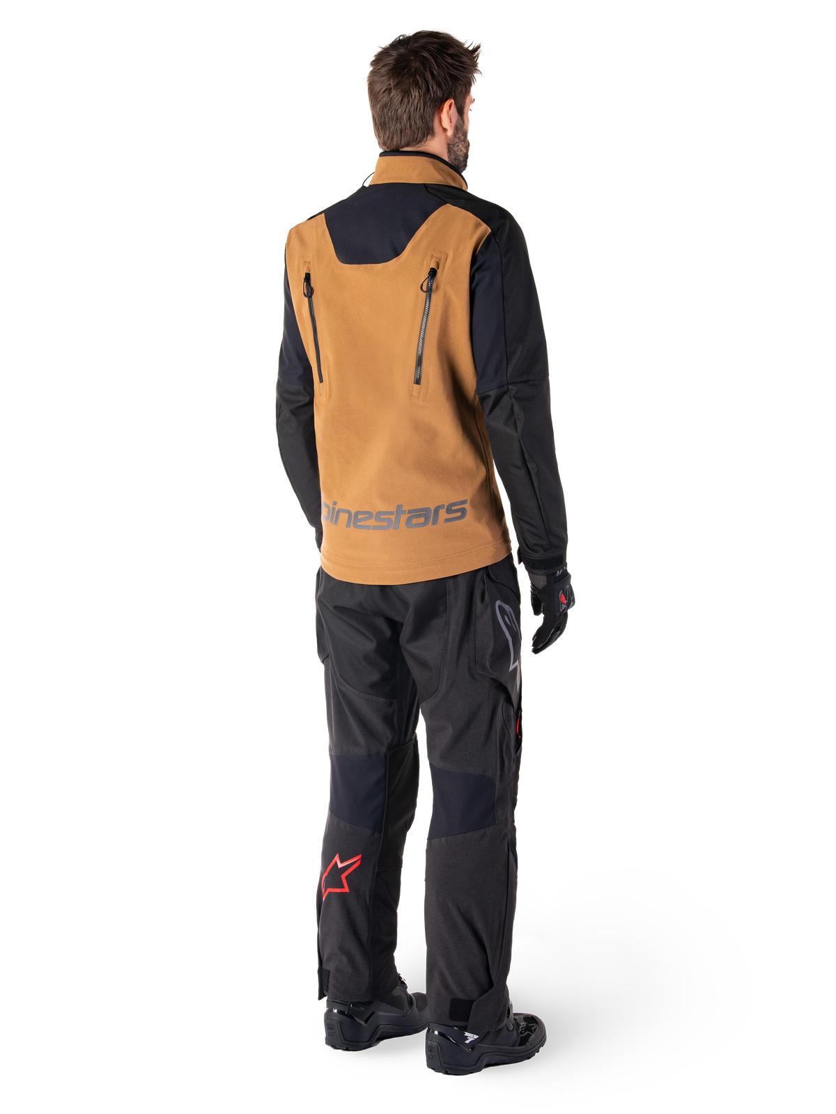 Alpinestars Hyde XT Stretch Drystar®XF jas en Hyde XT Drystar® broek, achteraanzicht van een rijder in zandkleurige en zwarte adventure touring uitrusting, jas met verticale rugritsen en Alpinestars-branding, zwarte broek met rood sterlogo op de kuit, gecombineerd met zwarte handschoenen en laarzen.