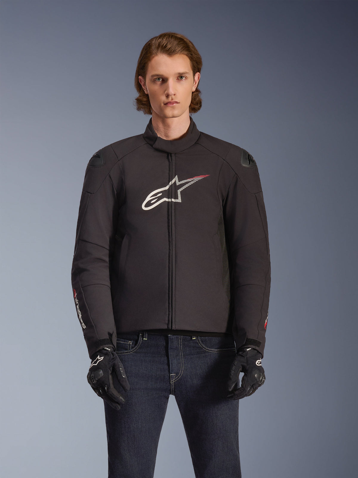 Alpinestars SMX Waterproof Jacket, zwarte textielen motorjas met wit en rood sterlogo op de borst, gedragen door een model met zwarte handschoenen en blauwe jeans, voorzien van externe schoudersliders en een sportieve pasvorm.