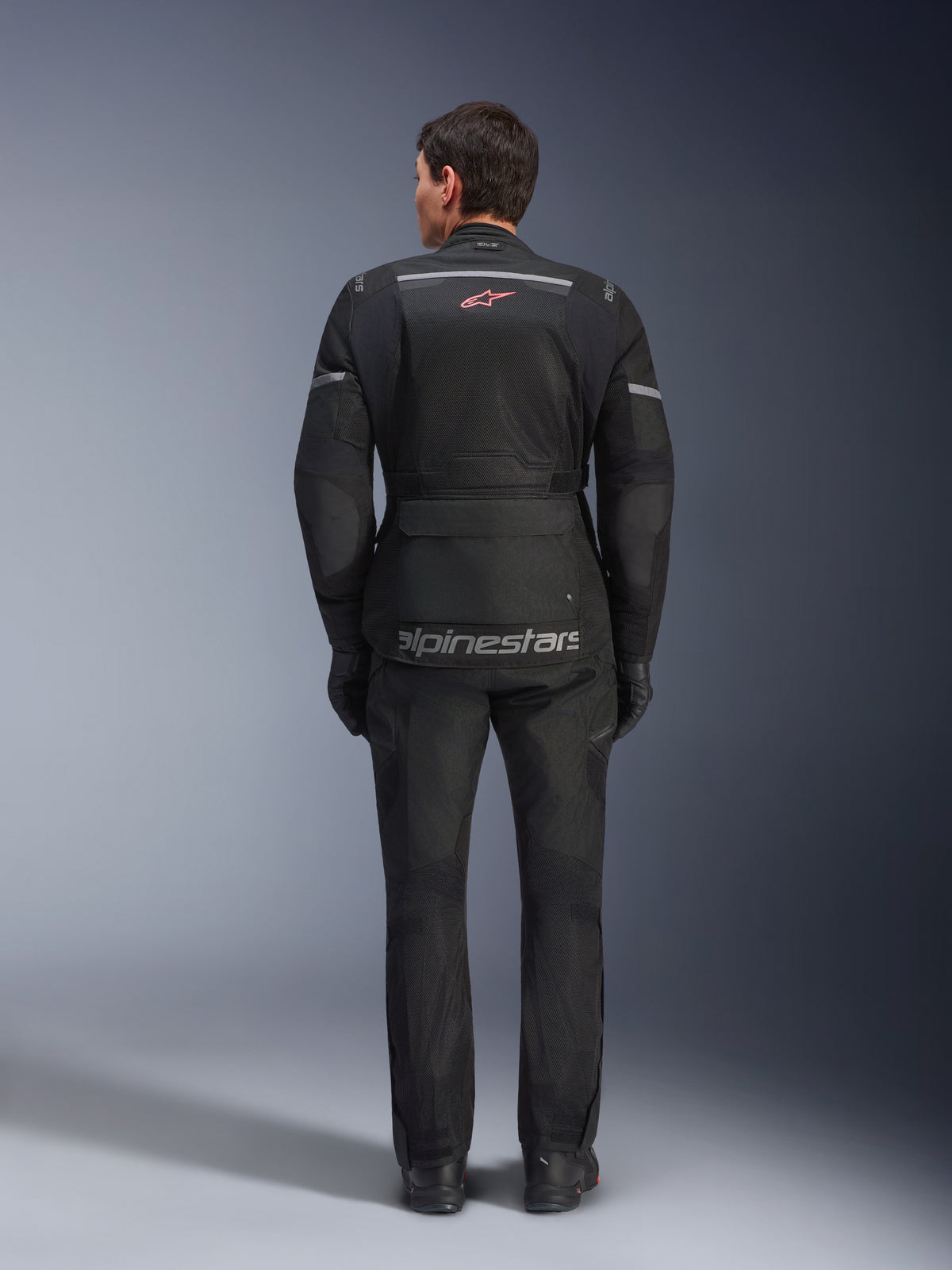 Achteraanzicht van een rijder die de Alpinestars Andes Air Drystar® Jas in het zwart draagt, voorzien van uitgebreide mesh ventilatiepanelen op de rug, een rood Alpinestars logo en reflecterende accenten, gecombineerd met een bijpassende motorbroek en handschoenen voor adventure touring.