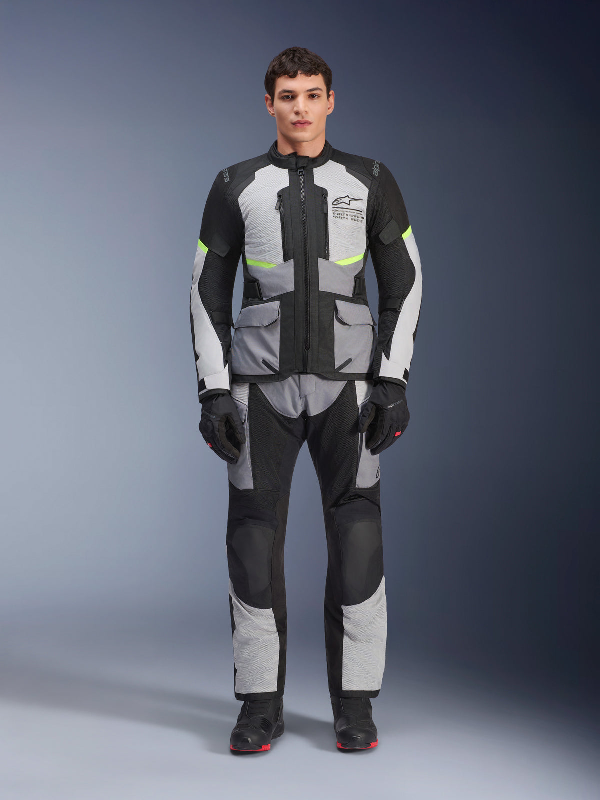 Alpinestars Andes Air Drystar® jas in Ice Gray, Dark Gray en Black, gedragen door een mannelijk model met bijpassende motorbroek en handschoenen, adventure touring ontwerp met grote zakken aan de voorzijde en fluorescerend gele accenten, studio vooraanzicht