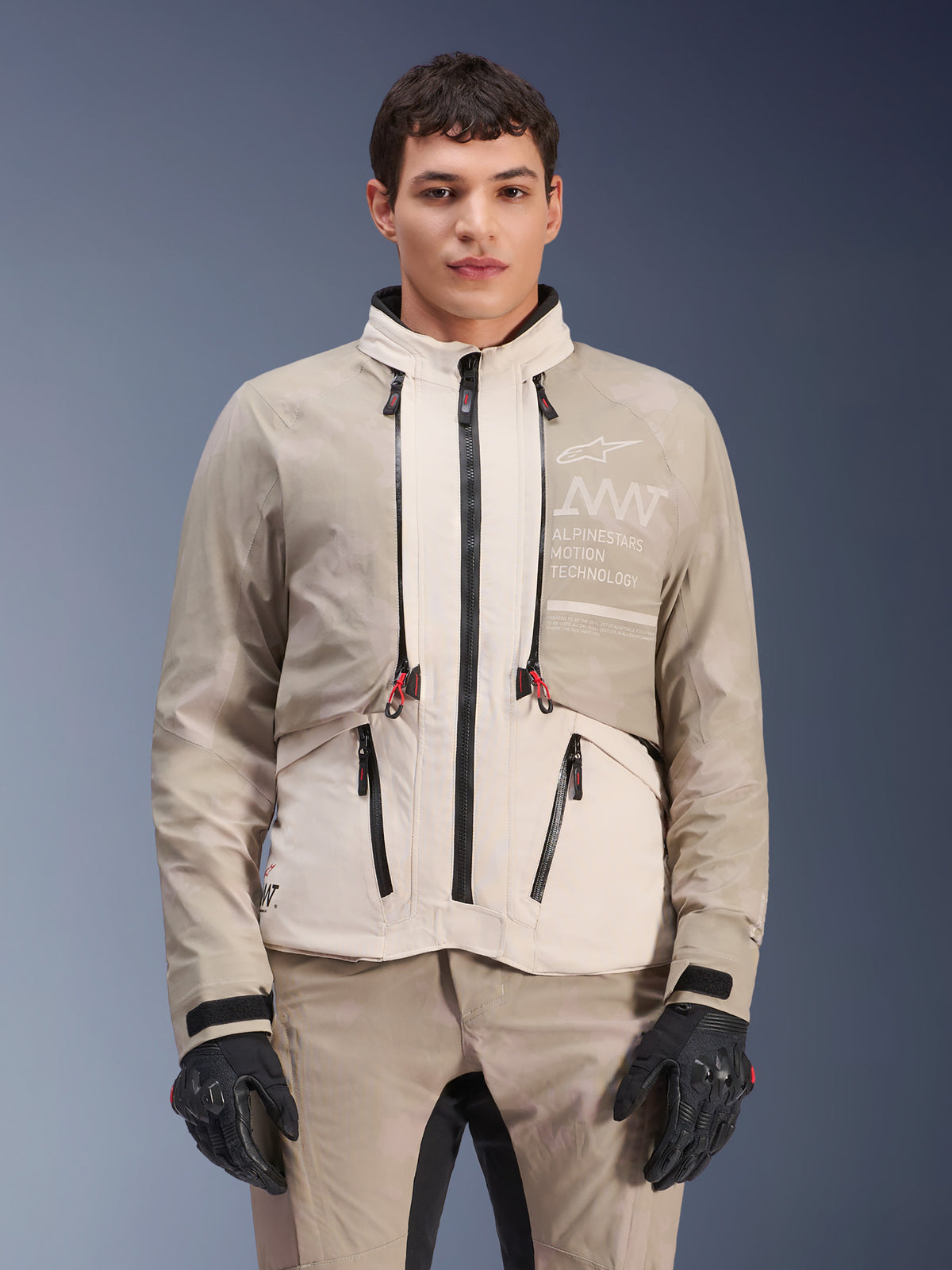 Alpinestars AMT-10LAB Drystar® XF jas, Adventure motorjas, White Sand Aluminum Camo, modulair ontwerp met zwarte ritsen en rode accenten, gedragen met bijpassende broek en zwarte handschoenen, voorzien van Alpinestars Motion Technology-branding op de borst, professioneel all-weather motorpak