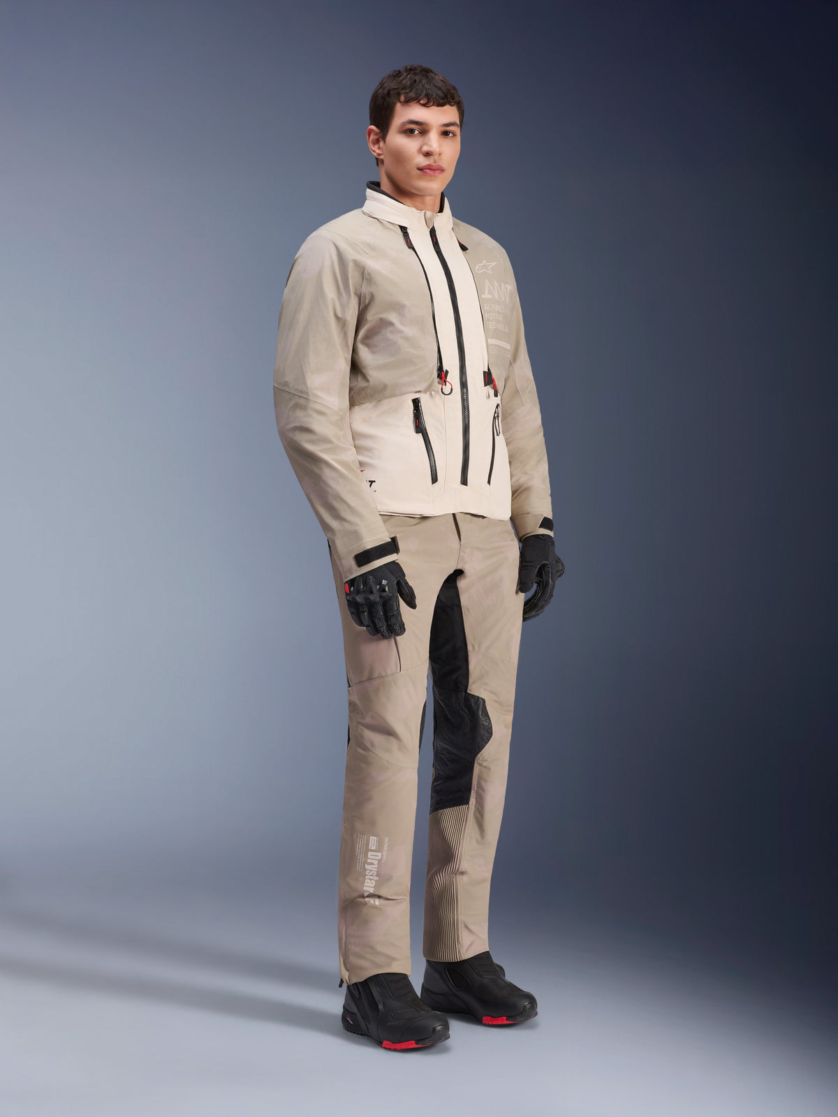 Alpinestars AMT-10LAB Drystar® XF jas, adventure motorjas, White Sand Aluminum Camo, gedragen met bijpassende adventure broek, zwarte motorhandschoenen en laarzen, studio-opname van het volledige lichaam, beschermende motorkleding voor alle seizoenen