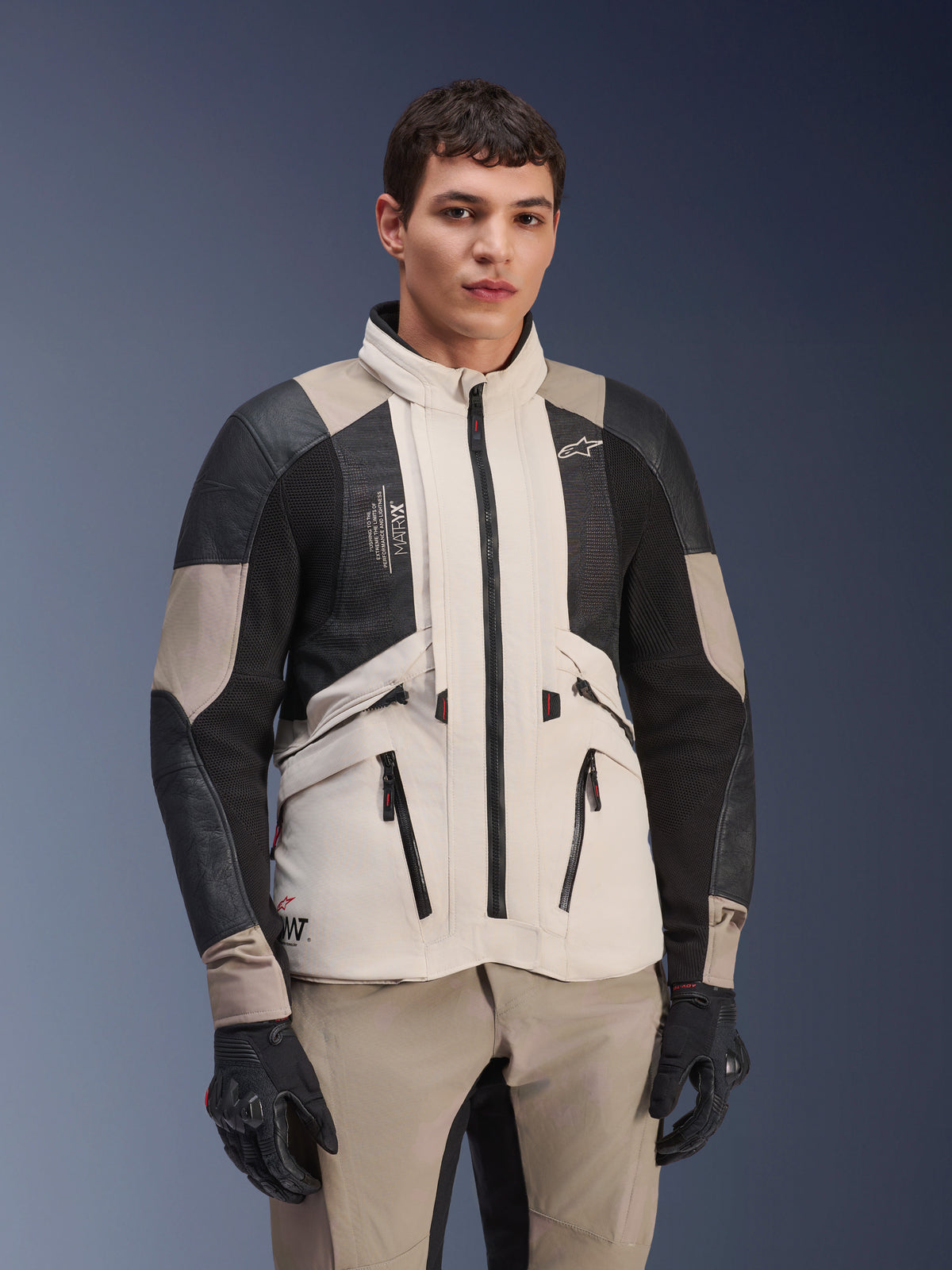 Alpinestars AMT-10LAB Drystar® XF jas, White Sand Aluminum Camo, adventure motorjas met zwarte MATRYX® mesh panelen en lederen verstevigingen, gedragen door een model met zwarte handschoenen en bijpassende zandkleurige broek, studioportret