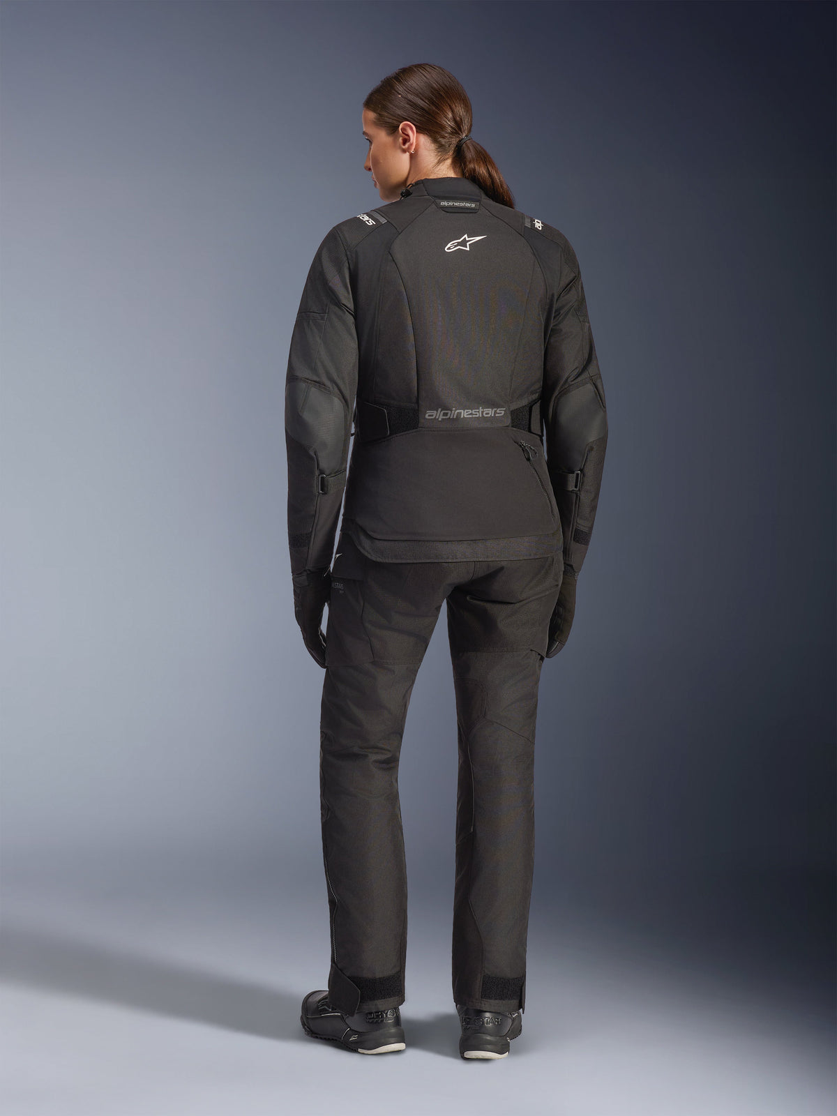 Vrouwelijke motorrijder van achteren gezien in de Alpinestars Stella Andes V4 Drystar® jas, zwart, gecombineerd met een bijpassende motorbroek, wit Astar-logo op de rug, verlengde achterzoom, complete adventure-touring motoroutfit inclusief zwarte handschoenen en laarzen