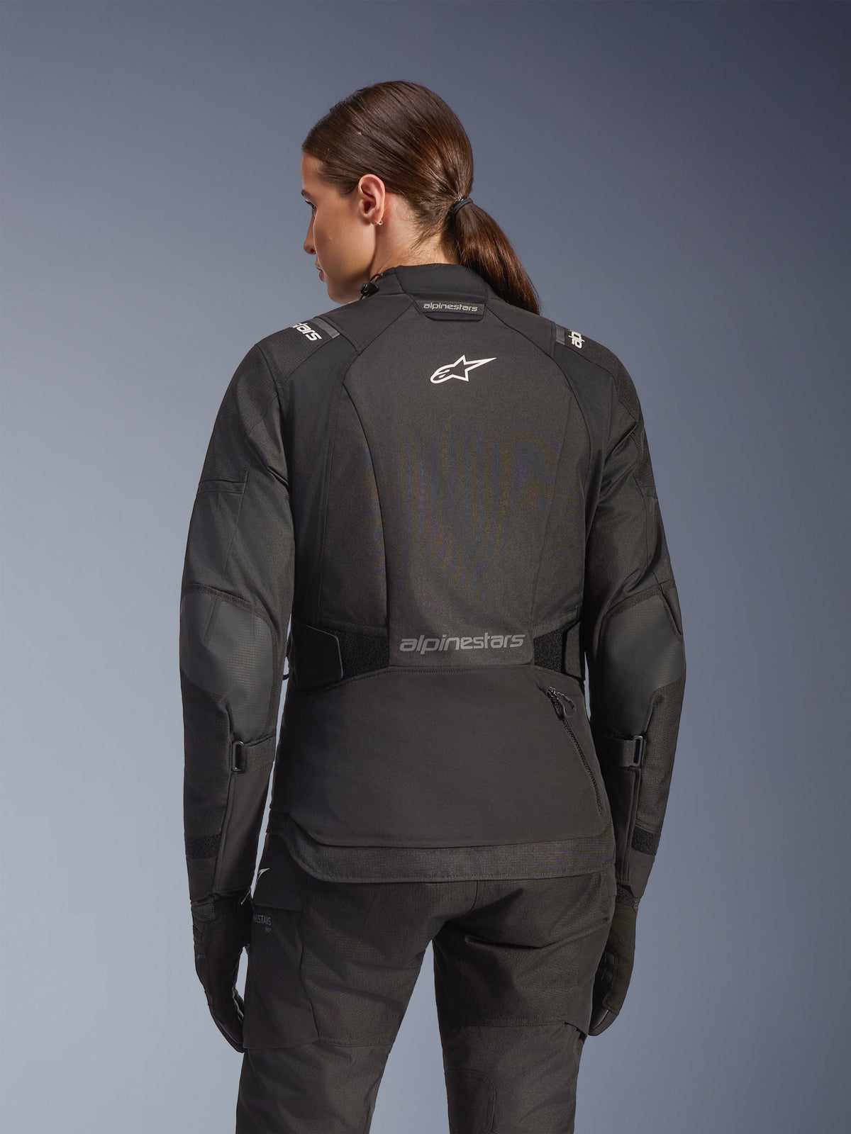 Vrouw in Alpinestars Stella Andes V4 Drystar jas in het zwart, achteraanzicht met wit logo op de rug en verstevigde elleboogpanelen, gecombineerd met bijpassende zwarte broek en handschoenen, adventure touring motoroutfit