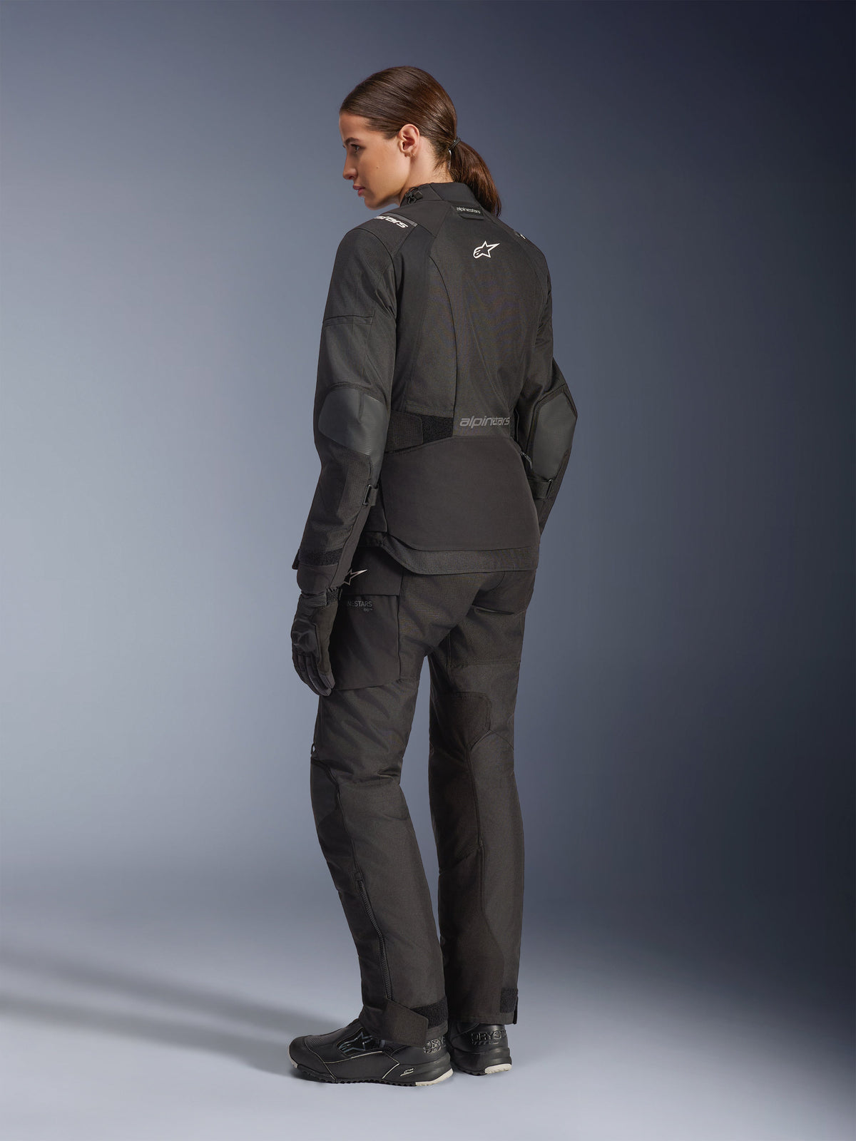 Alpinestars Stella Andes V4 Drystar® jas, zwarte adventure motorjas, driekwart achteraanzicht, gecombineerd met bijpassende textiele broek en handschoenen, voorzien van witte Alpinestars-logo's op de rug en taille, versterkte beschermende panelen en een verlengde achterzoom, vrouwelijke motorrijder