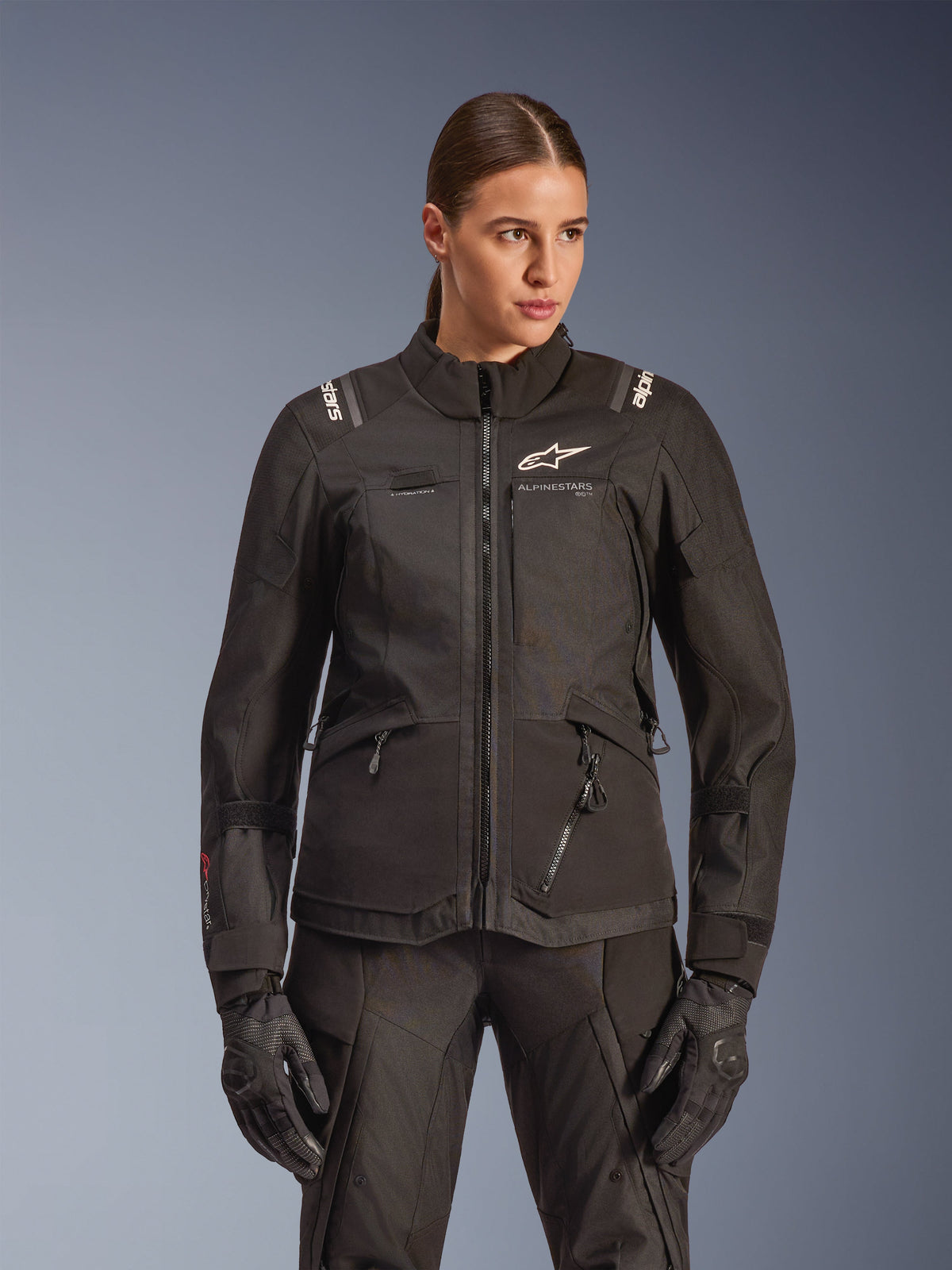 Alpinestars Stella Andes V4 Drystar motorjas, dames touring motorjas, zwart, gedragen door een model met bijpassende motorbroek en handschoenen, voorzien van een ritssluiting aan de voorzijde, wit logo op de borst en Alpinestars-branding op de schouders, stijlvolle all-weather uitrusting