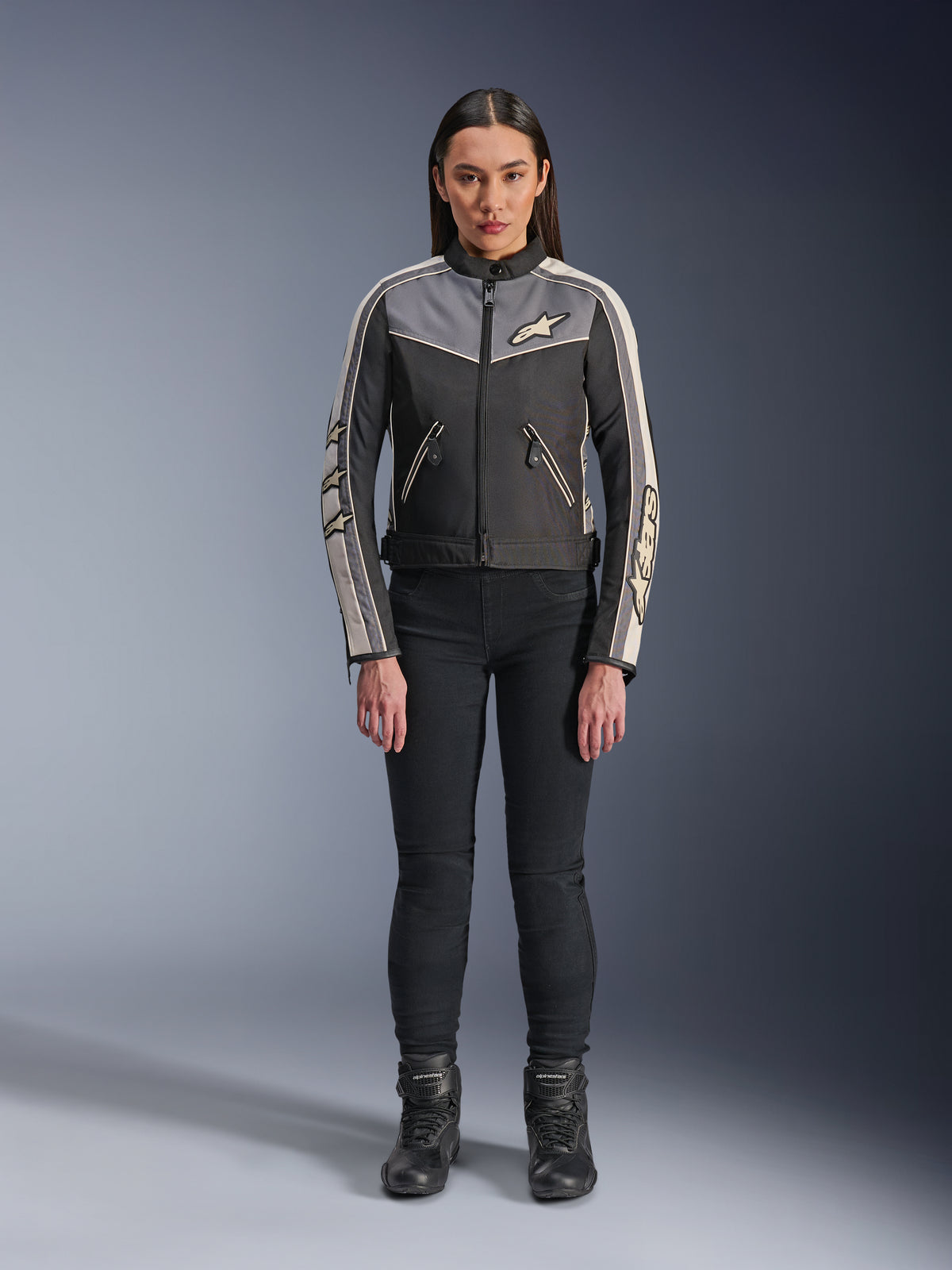 Vrouw in Alpinestars Stella T-Dyno WR-jas, zwart, donkergrijs en ecru, vooraanzicht, textiele motorjas met een op vintage geïnspireerd ontwerp, voorzien van merklogo's op de borst en mouwen, gedragen met een zwarte broek en laarzen