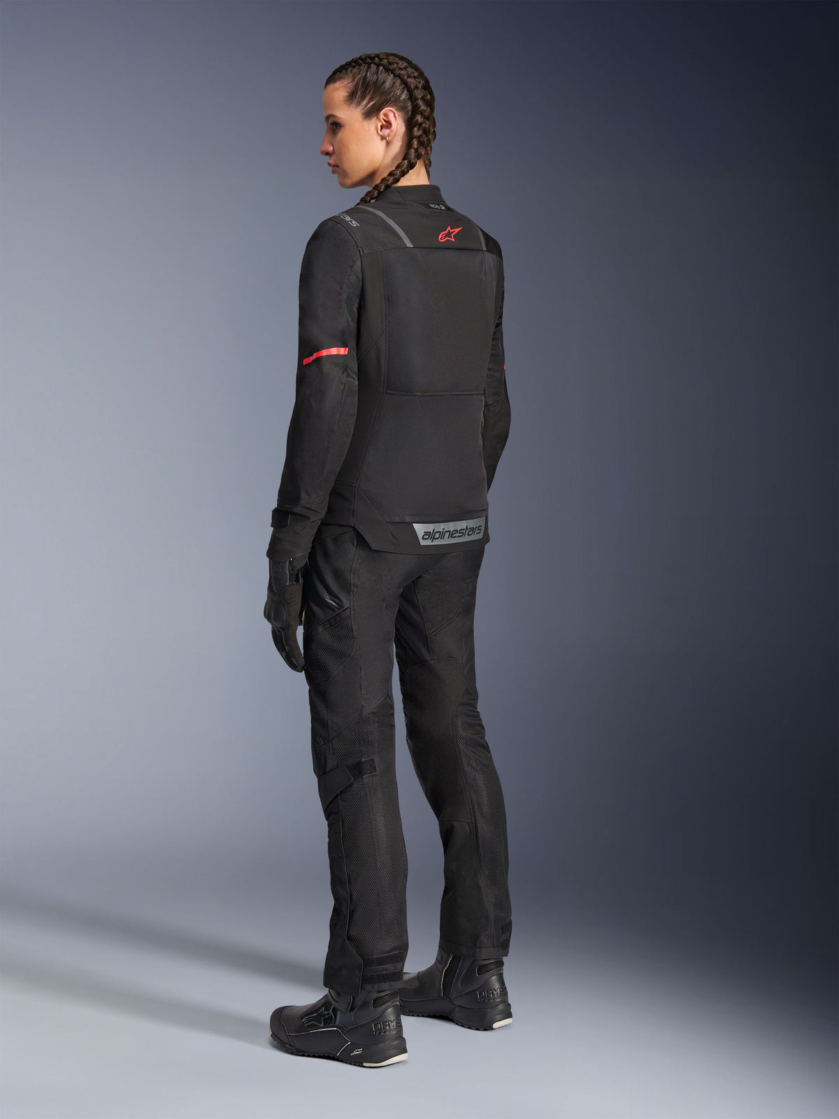 Vrouwelijke motorrijder in zwarte Alpinestars Stella Andes Air Drystar® broek en bijpassende Stella ST-2 Air jas, achteraanzicht toont de technische textielconstructie en meshpanelen, rood sterlogo op de rug van de jas, gecombineerd met zwarte motorlaarzen en handschoenen.