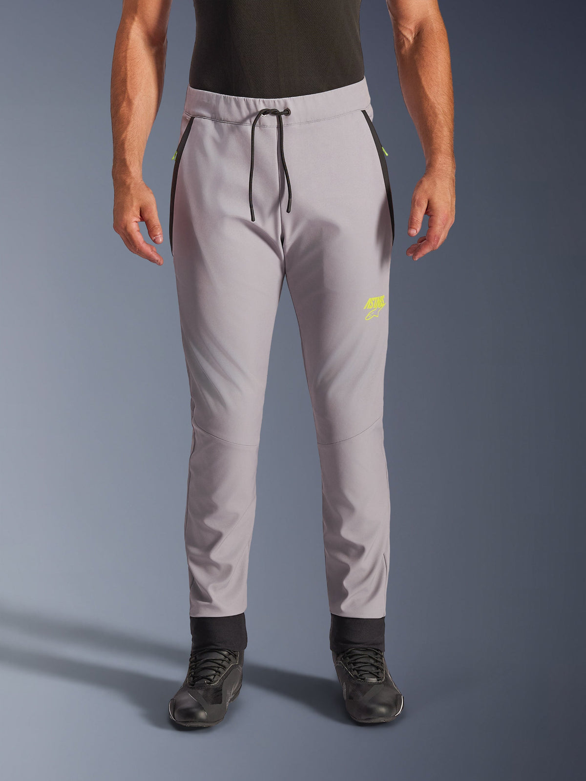 Alpinestars Aeroshell Pants, middengrijs en fluoriserend geel, crossover softshell motorbroek met een zwart trekkoord in de taille en aansluitende enkelmanchetten, geel logo op de linkerpijp, voorgevormde knieën en een sportief jogger-stijl silhouet