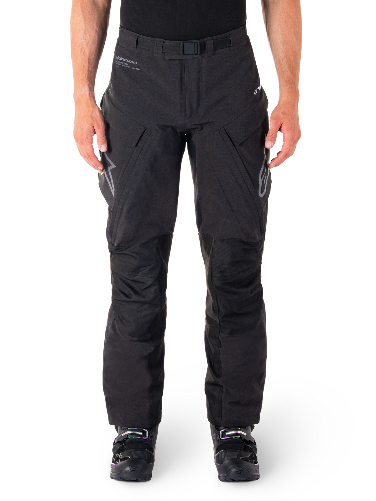 Alpinestars Hyde XT Drystar broek, zwarte adventure touring motorbroek, gedragen door een rijder met zwarte laarzen, voorzien van grijze logo's op de dijen, geïntegreerde riem, versterkte kniepanelen en waterdichte textielconstructie