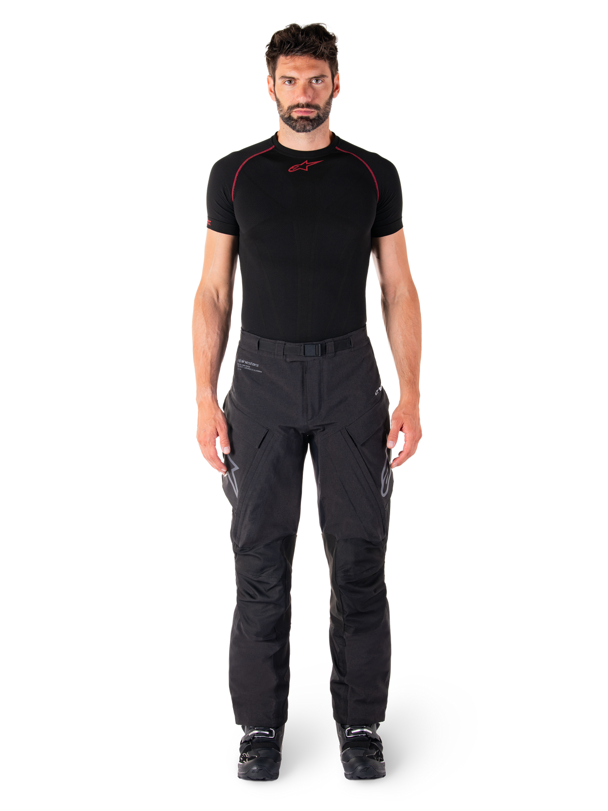 Alpinestars Hyde XT Drystar® Broek, adventure touring motorbroek, zwart, gedragen door een model met een zwarte basislaag met korte mouwen en zwarte laarzen, voorzien van technisch stretchmateriaal, versterkte kniepanelen en ergonomische cargozakken.