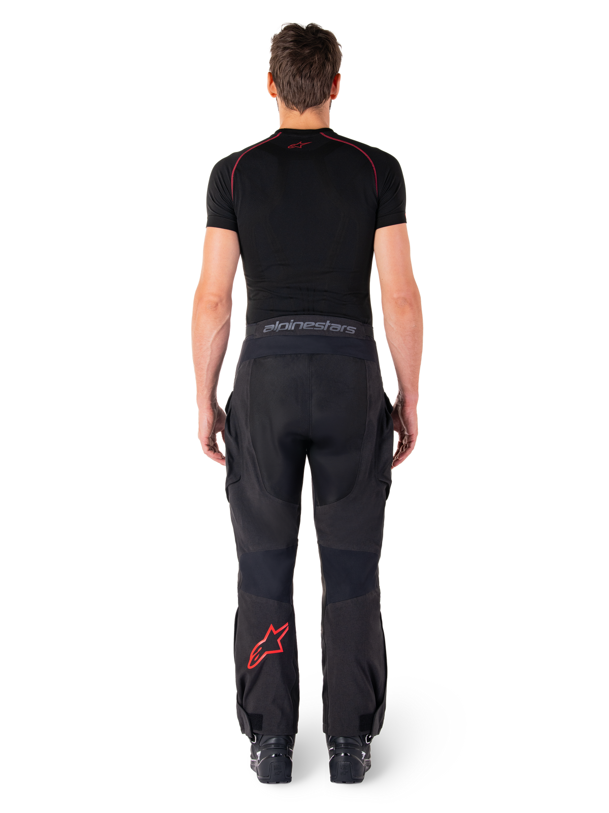 Alpinestars Hyde Xt Drystar® broek, adventure touring motorbroek, zwart en felrood, achteraanzicht van een rijder die de broek en een zwarte basislaag draagt, met een rood sterlogo op het linkerbeen en grijze merknaam op de tailleband.