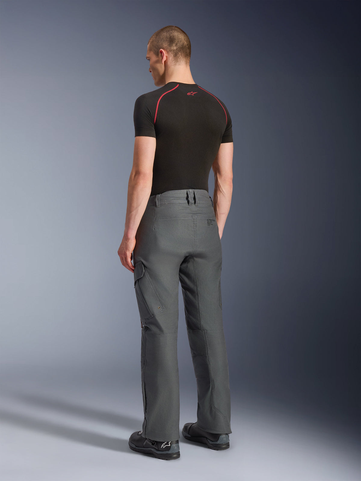 Alpinestars Borrego Drystar® broek, adventure motorbroek, antracietgrijs, achteraanzicht, voorzien van een gewaxte canvas-look met cargozakken, gecombineerd met een zwart t-shirt en motorlaarzen, duurzaam ontwerp voor weg- en adventure-ritten
