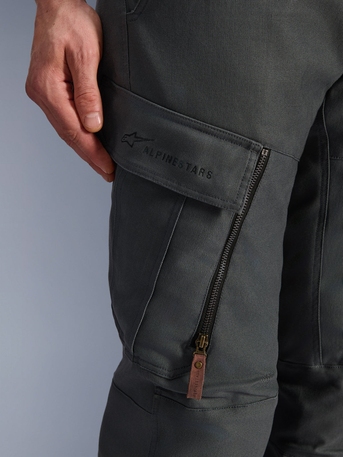 Alpinestars Borrego Drystar® broek, antraciet grijs, close-up van de cargozak op het dijbeen met geborduurd logo, getextureerd gewaxt canvas materiaal, verticale ventilatierits met een bruine lederen ritstrekker