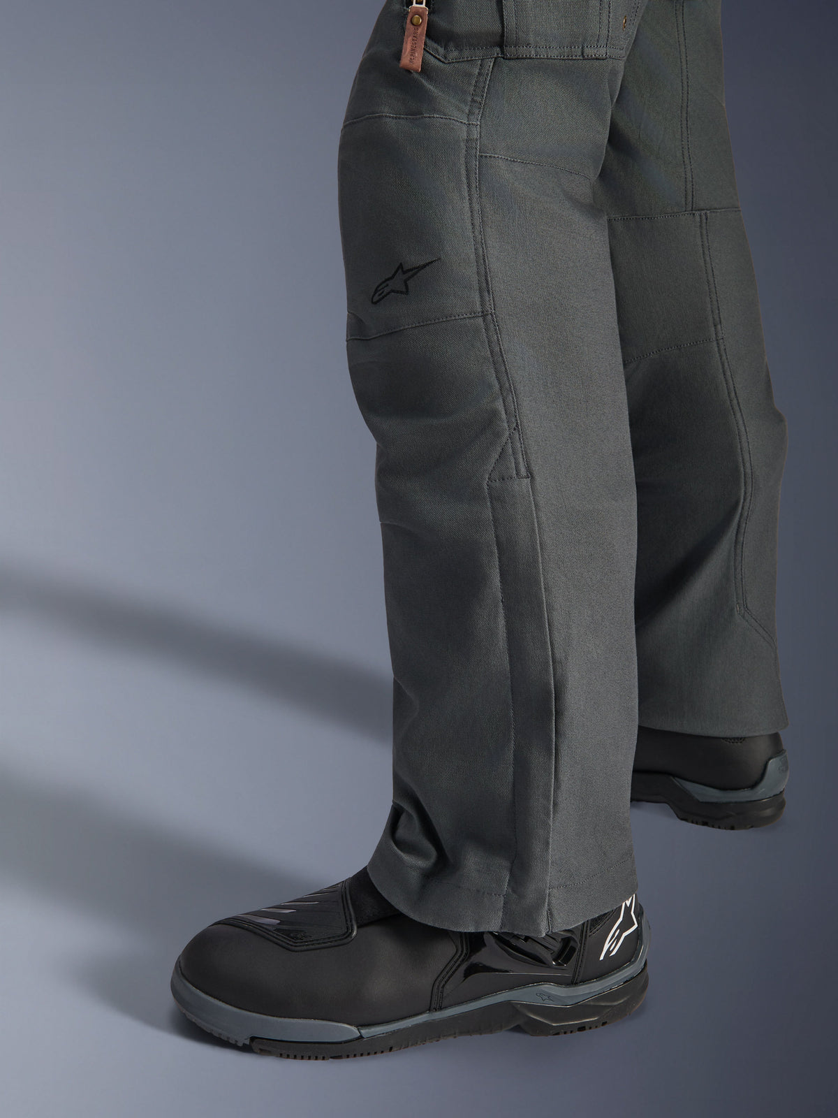 Alpinestars Borrego Drystar® broek, antracietgrijze adventure motorbroek met een gewaxt canvas uiterlijk, textielconstructie van opzij gezien met zwarte motorlaarzen, robuust ontwerp voor weg- en avontuurlijk toeren