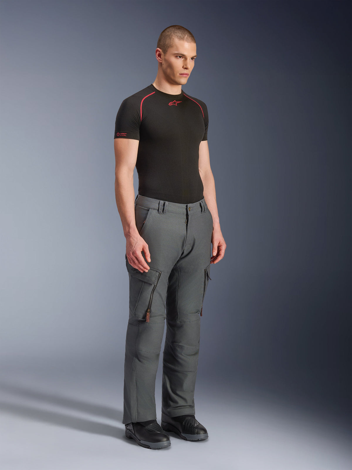 Mannelijk model draagt Alpinestars Borrego Drystar® broek, antracietgrijze adventure motorbroek, op werkkleding geïnspireerd ontwerp met cargo-zakken en ventilatieritsen, gecombineerd met een zwarte basislaag met korte mouwen en zwarte laarzen