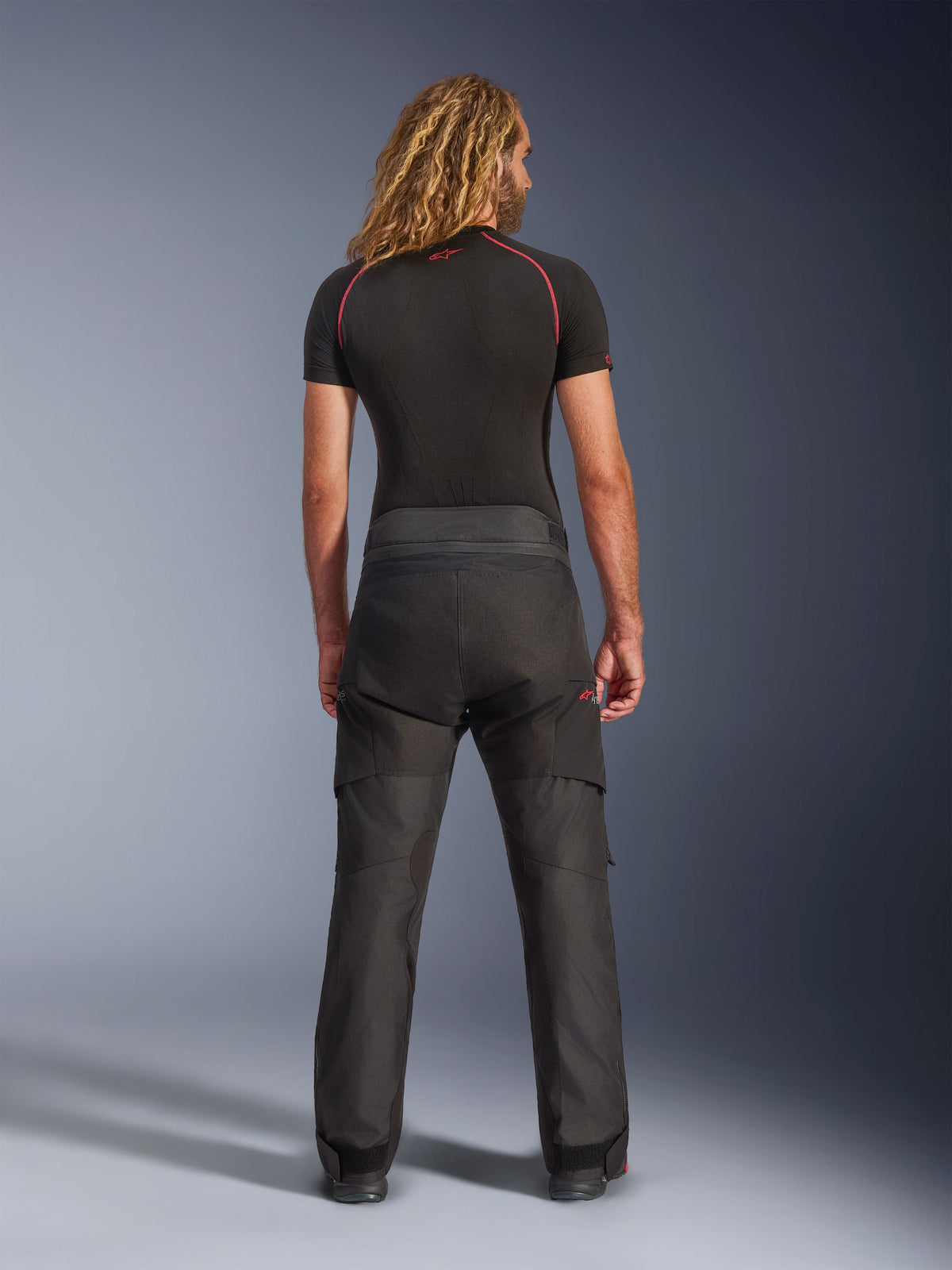 Rear view of a model wearing Alpinestars Andes V4 Drystar® Pants - Short, black adventure motorcycle pants, paired with a black base layer featuring red stitching, showcasing the durable textile construction and ergonomic fit.