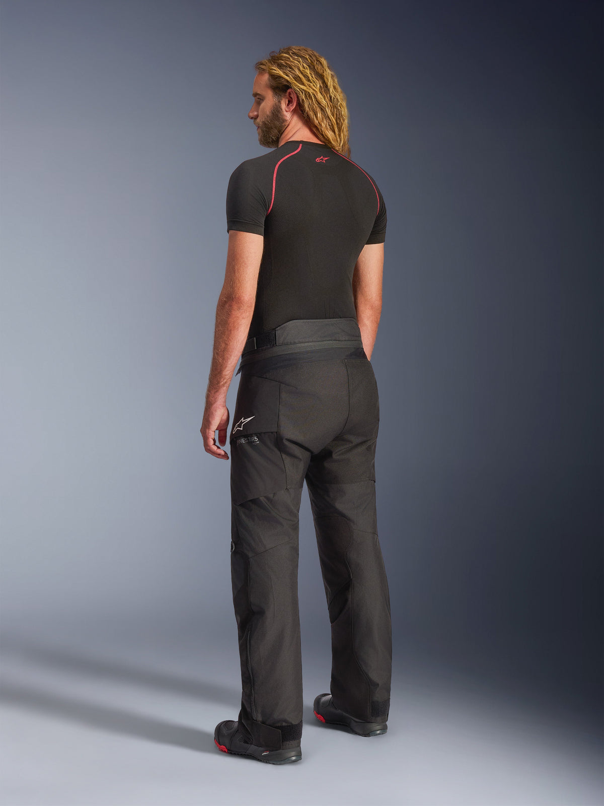 Alpinestars Andes V4 Drystar® Pants - Short, Black, back three-quarter view on a model with long blonde hair, worn with a black base layer tee with red stitching and motorcycle boots, technical adventure touring design with durable textile panels.