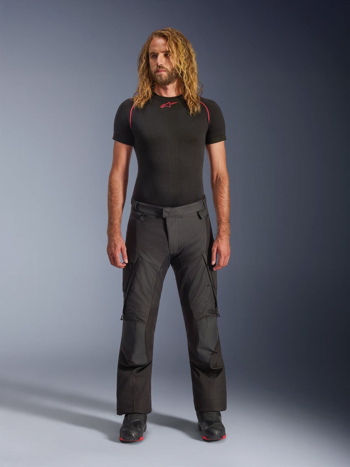 Male model with long hair wearing Alpinestars Andes V4 Drystar® Pants in Black, adventure touring motorcycle pants with reinforced knee panels and cargo pockets, styled with a black short-sleeve base layer and riding boots.