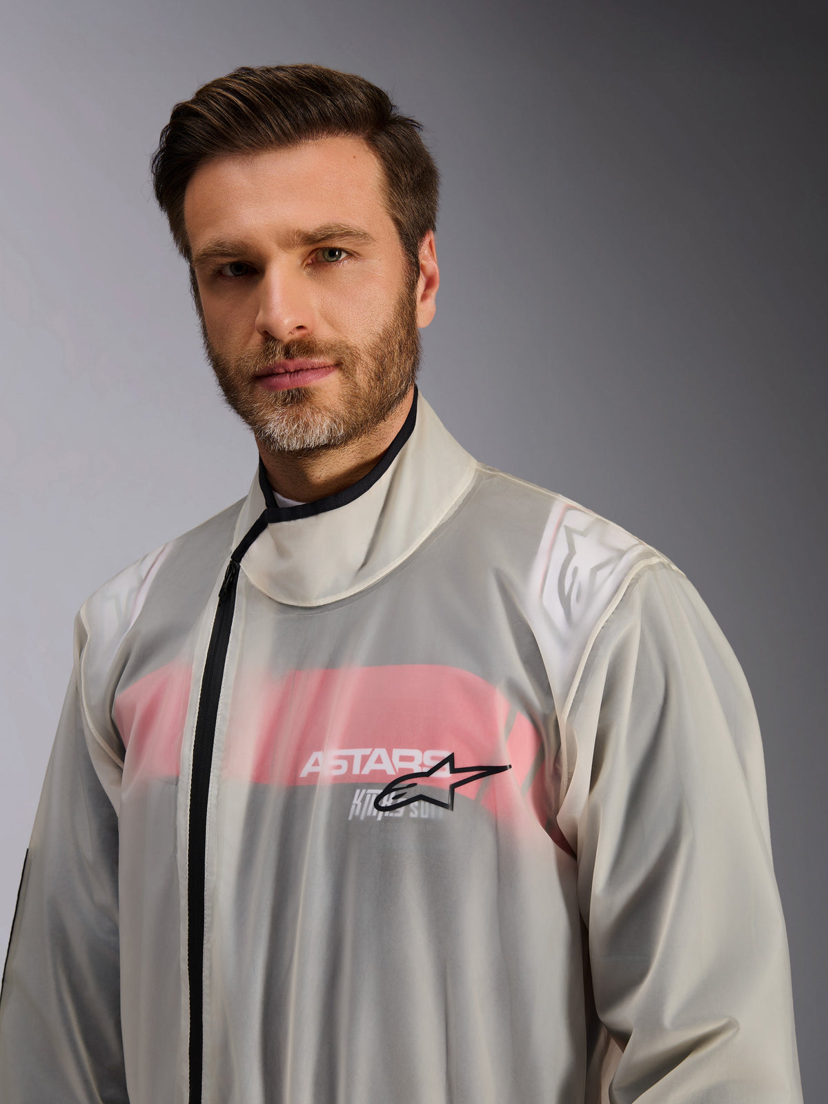 Alpinestars Kart Regenpak, karting regenoverpak, transparant, gedragen door een man, doorzichtig waterdicht materiaal waaronder details van een rood-wit racepak zichtbaar zijn, hoge kraag met zwarte ritssluiting en Alpinestars sterlogo op de borst