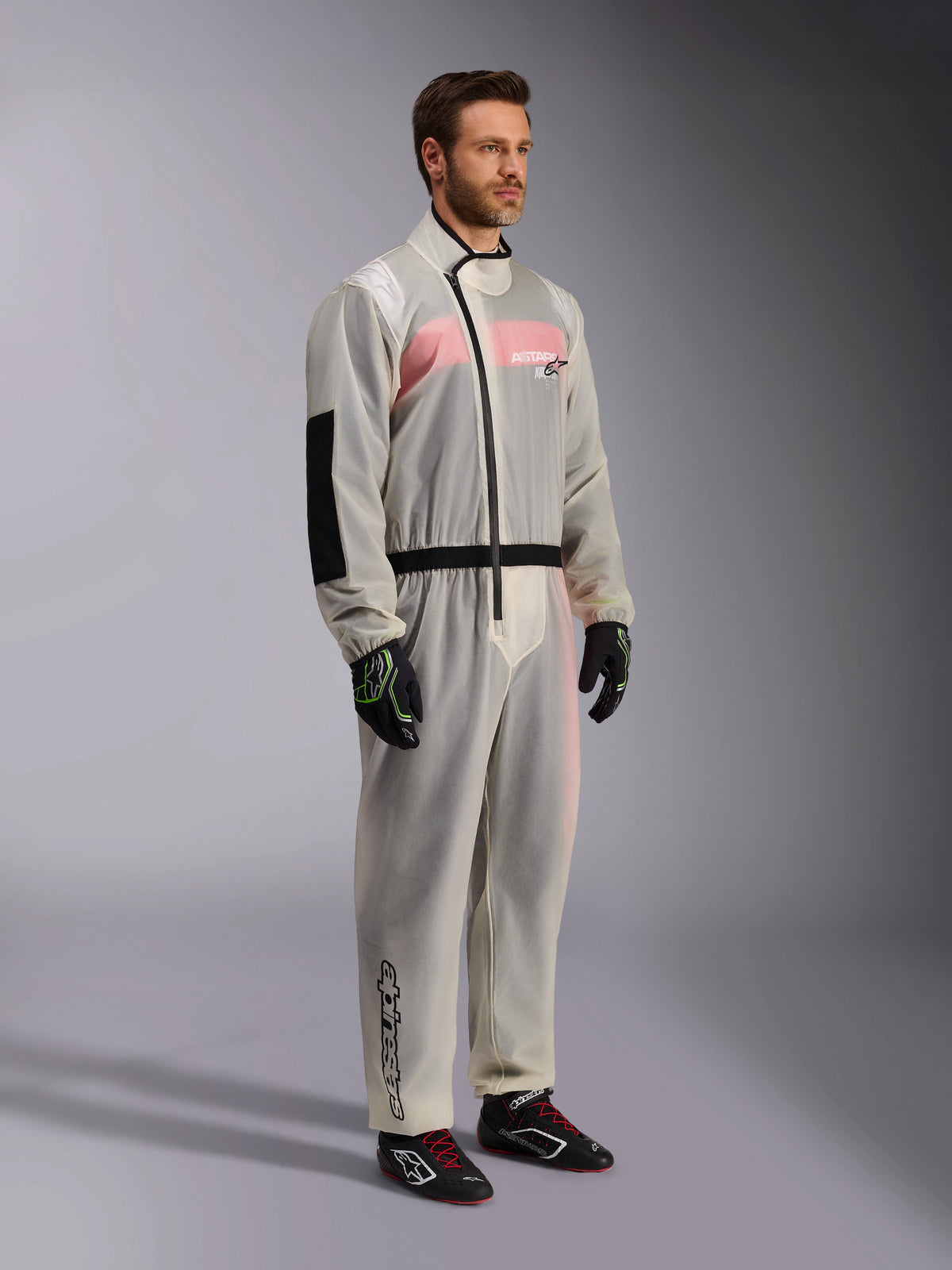 Alpinestars Kart Regenpak, transparant waterdicht karting overpak, semi-transparant materiaal dat een roze en wit racepak eronder toont, voorzien van een zwarte asymmetrische ritssluiting, versterkt elleboogstuk en zwarte tailleband, gedragen met zwarte karthandschoenen en raceschoenen.
