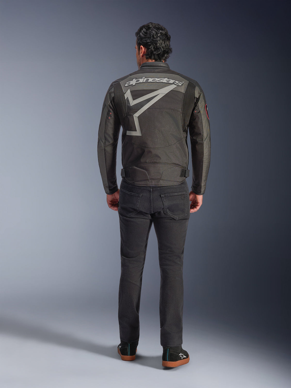 Alpinestars Axton Jacket, motorjas, Black Anthracite Red Tangerine, achteraanzicht met groot grijs Alpinestars-logo en stergrafiek, accordeonpanelen op de schouders, gedragen met zwarte broek en casual zwarte schoenen