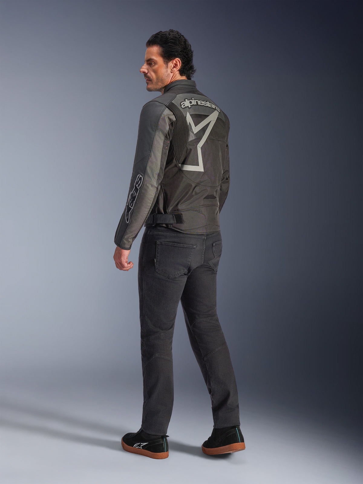 Alpinestars Axton Jacket, motorjas voor heren, zwart en antraciet, achteraanzicht met een groot grijs rondom lopend logo en accordeonpanelen op de schouders, gedragen met een donkere motorbroek en zwarte sneakers, urban contemporary stijl met race-erfgoed.