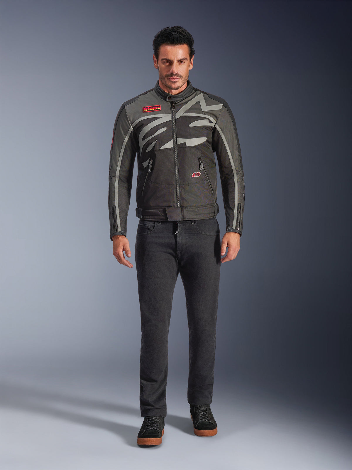 Alpinestars Axton jas, motorjas, Black Anthracite Red Tangerine, gedragen door een model met een donkere broek, voorzien van een rondom lopend grijs Alpinestars-logo, rode merkpatches en een waterbestendige textiele buitenlaag voor stadsritten