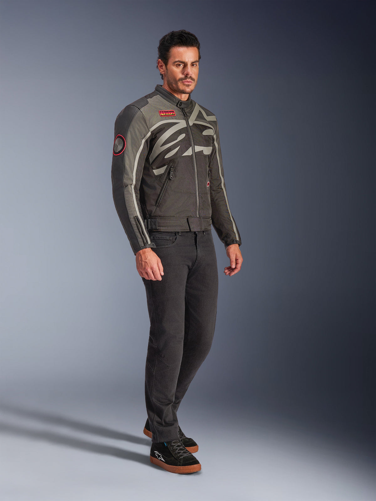 Mannelijk model draagt de Alpinestars Axton Jas in Black Anthracite Red Tangerine, vintage-geïnspireerde motorjas met een rondom lopend logo en rode accenten, gecombineerd met een zwarte broek en zwarte motorsneakers, urban contemporary motorkleding