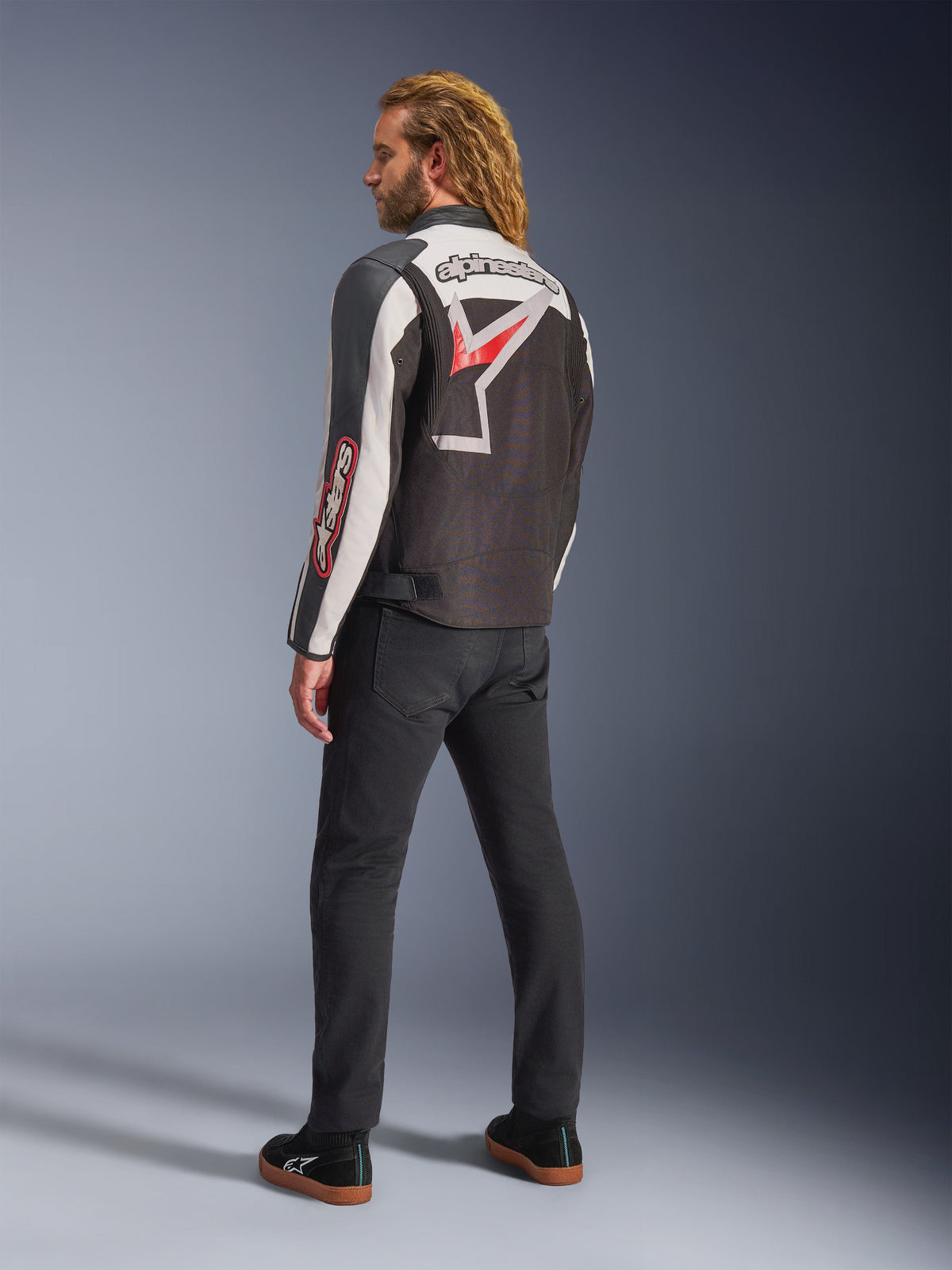 Alpinestars Axton Jas, motorjas, zwart, gebroken wit en felrood, achteraanzicht gedragen door mannelijk model, voorzien van groot Alpinestars-sterlogo en woordmerk op de rug, vintage racing-geïnspireerd ontwerp met accordeonpanelen, gecombineerd met zwarte broek en sneakers