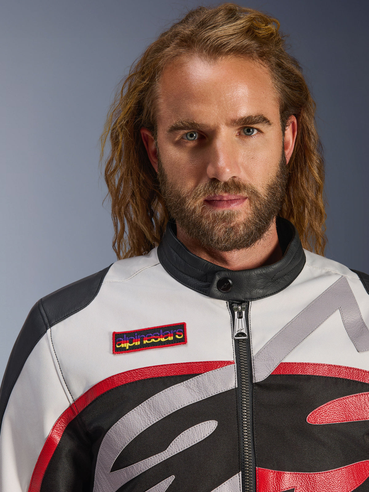 Man in de Alpinestars Axton Jacket, vintage-geïnspireerde motorjas in Black Off White en Bright Red, close-up weergave van de merkpatch op de borst en de minimalistische biker-kraag, eigentijdse motorkleding voor op de weg