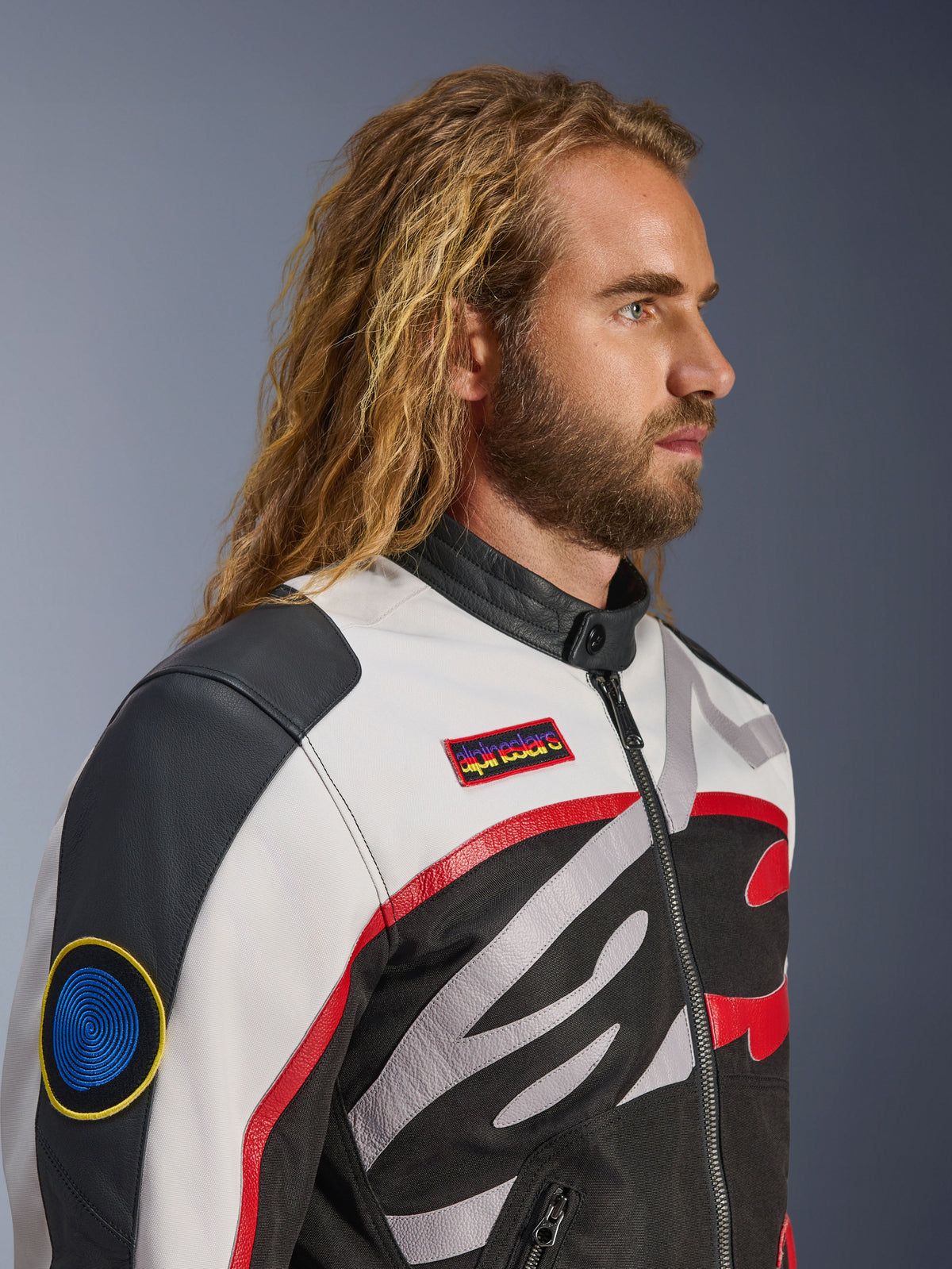 Man met lang haar en baard draagt het Alpinestars Axton Jacket, motorjas, zwart, off-white en felrood, vintage racing-geïnspireerd ontwerp met merkpatches en rondom lopend logo, close-up profielaanzicht, urban contemporary stijl