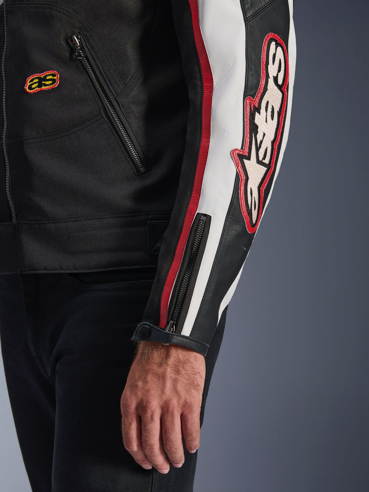 Alpinestars Axton Jas, close-up van mouw en torso, zwart met gebroken wit en felrode accenten, groot geborduurd Alpinestars-logo op de mouw, verticale racestrepen, manchet met ritssluiting en drukknoop, robuuste textielconstructie.