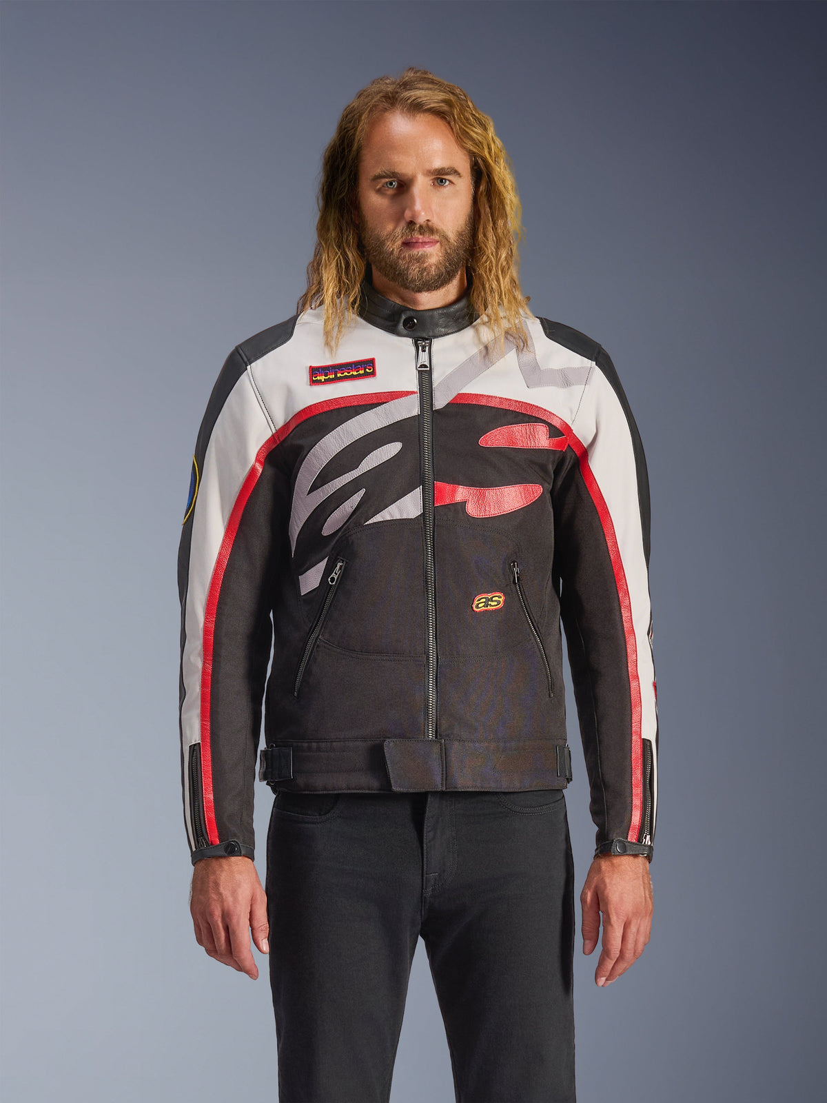 Alpinestars Axton Jas, motorjas, zwart, off-white en felrood, voorzien van opvallende abstracte patronen op de borst en een rondom lopend logo, mouwen met rode strepen, vintage-geïnspireerd ontwerp met een minimalistische kraag, gedragen door een model met zwarte jeans.
