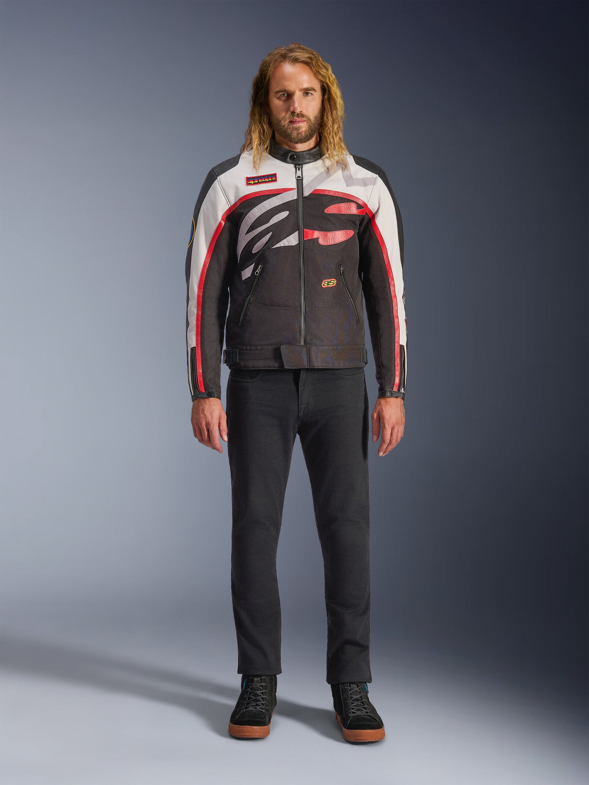 Alpinestars Axton Jacket, motorjas, zwart, gebroken wit en felrood, gedragen door een model met een zwarte broek en sneakers, voorzien van een opvallend rondom lopend logo en een vintage op de racerij geïnspireerd ontwerp