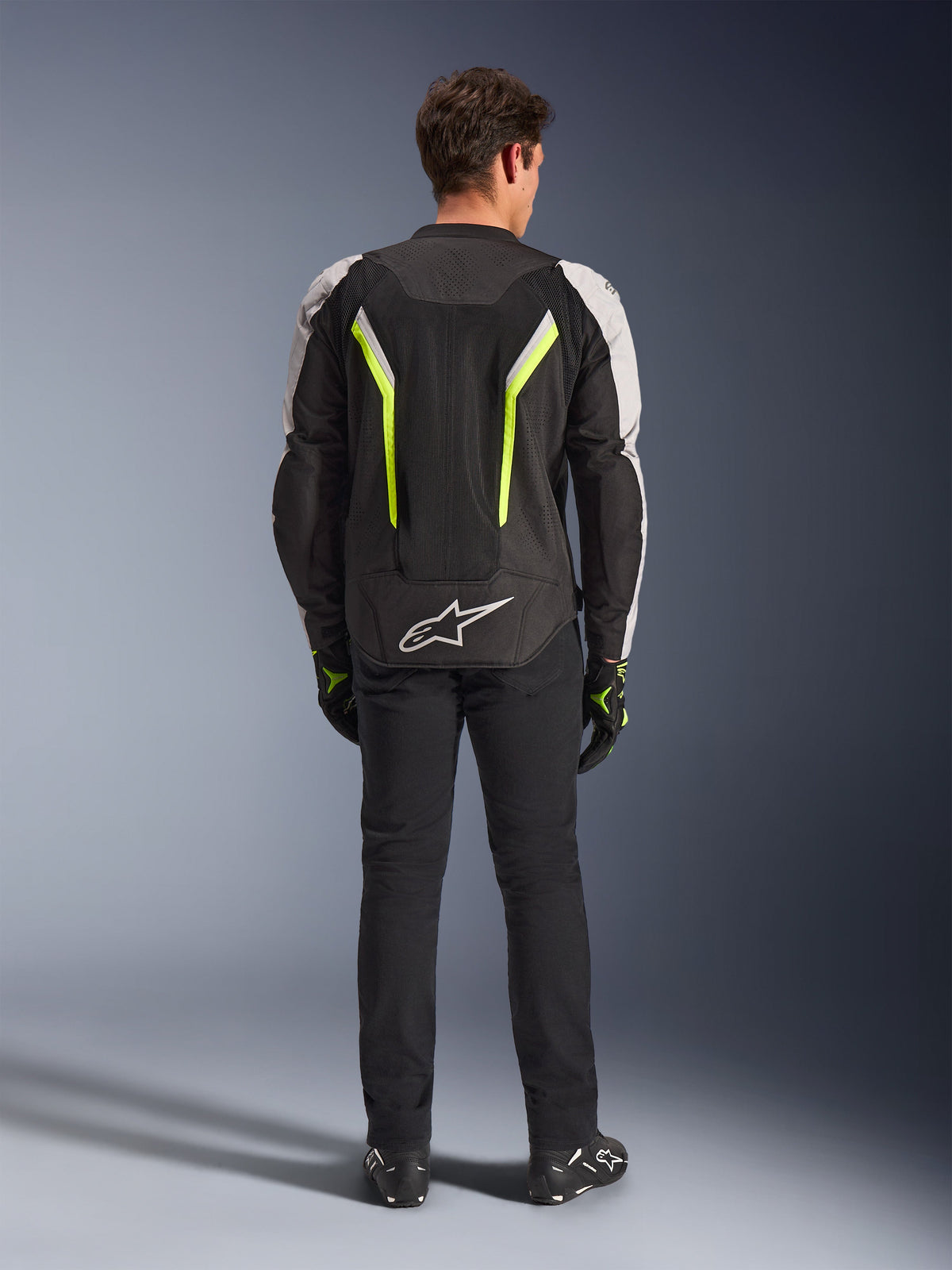 Alpinestars AST V3 Air Jacket, textiel motorjas, zwart, zilver en fluogeel, achteraanzicht met mesh ventilatiepanelen en fluorescerende accenten, gedragen met zwarte broek en motorhandschoenen, sportieve motoruitrusting